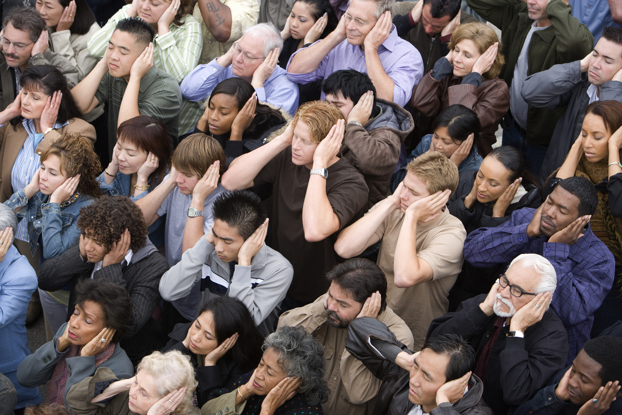 A large group of diverse people standing closely together outdoors, covering their ears with both hands. The individuals have serious expressions, and the background is densely packed with people.