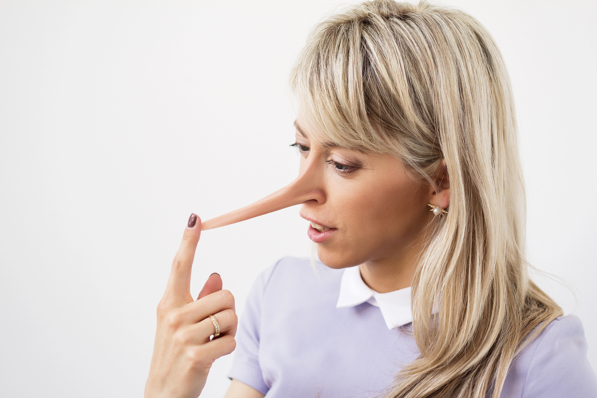 A woman with long blonde hair and wearing a light purple top and a white collar points to her elongated nose, resembling Pinocchio's nose, while looking at it. The background is plain white.