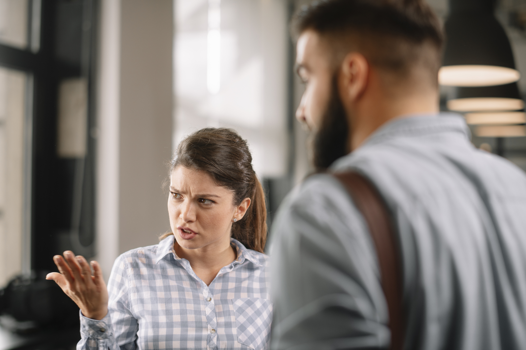 A woman with dark hair in a ponytail, wearing a blue and white checkered shirt, appears frustrated and gestures with her hand. A man with a beard, dressed in a light blue shirt and carrying a shoulder bag, stands facing her, partially out of focus.