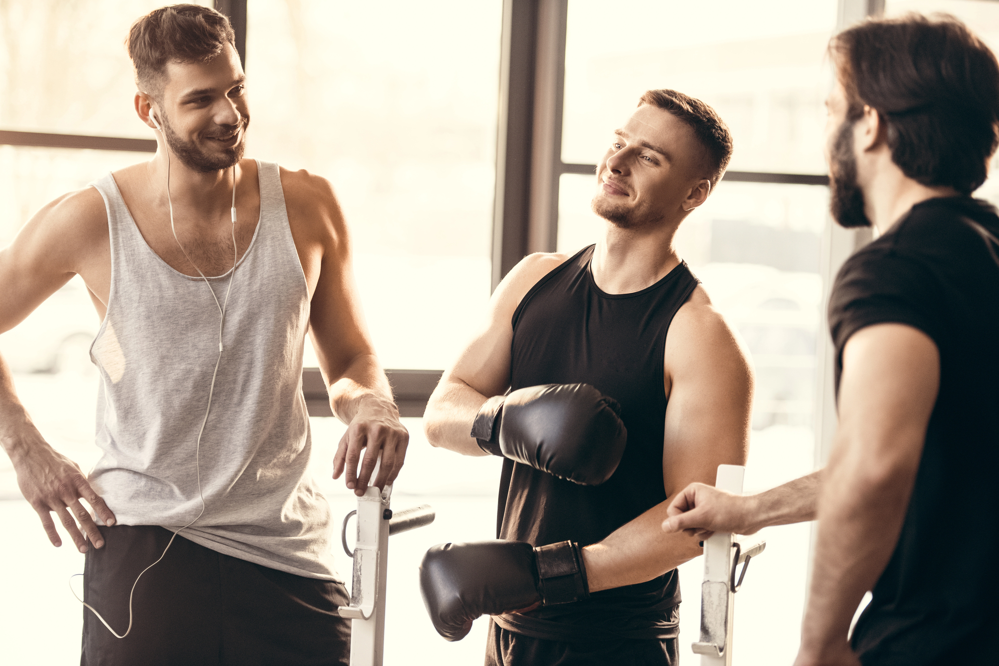 Three men are in a gym. One is wearing boxing gloves and a sleeveless shirt; the other two are casually dressed. They are standing near workout equipment and appear to be having a friendly conversation.