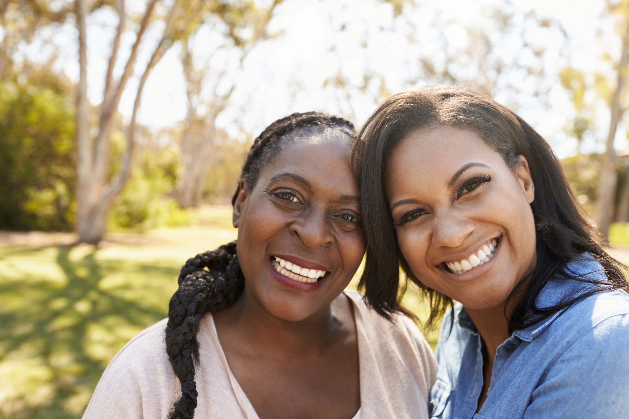 Two women are smiling brightly while standing close together outdoors. The woman on the left has long braided hair and the woman on the right has shoulder-length hair. They appear to be enjoying a sunny day in a park with trees in the background.