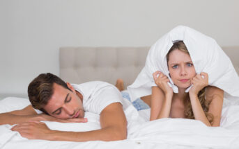 A man is sleeping peacefully on a bed while a woman next to him looks frustrated and covers her ears with a pillow. The woman appears to be disturbed, possibly by noise, as she looks upset. The scene is set in a well-lit bedroom with white bedding.