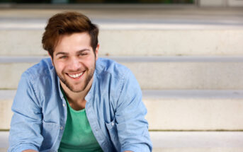 A smiling man with a beard and short, brown hair is sitting on a set of light-colored concrete steps. He is wearing a light blue, button-up shirt over a green t-shirt. The background is slightly blurred.