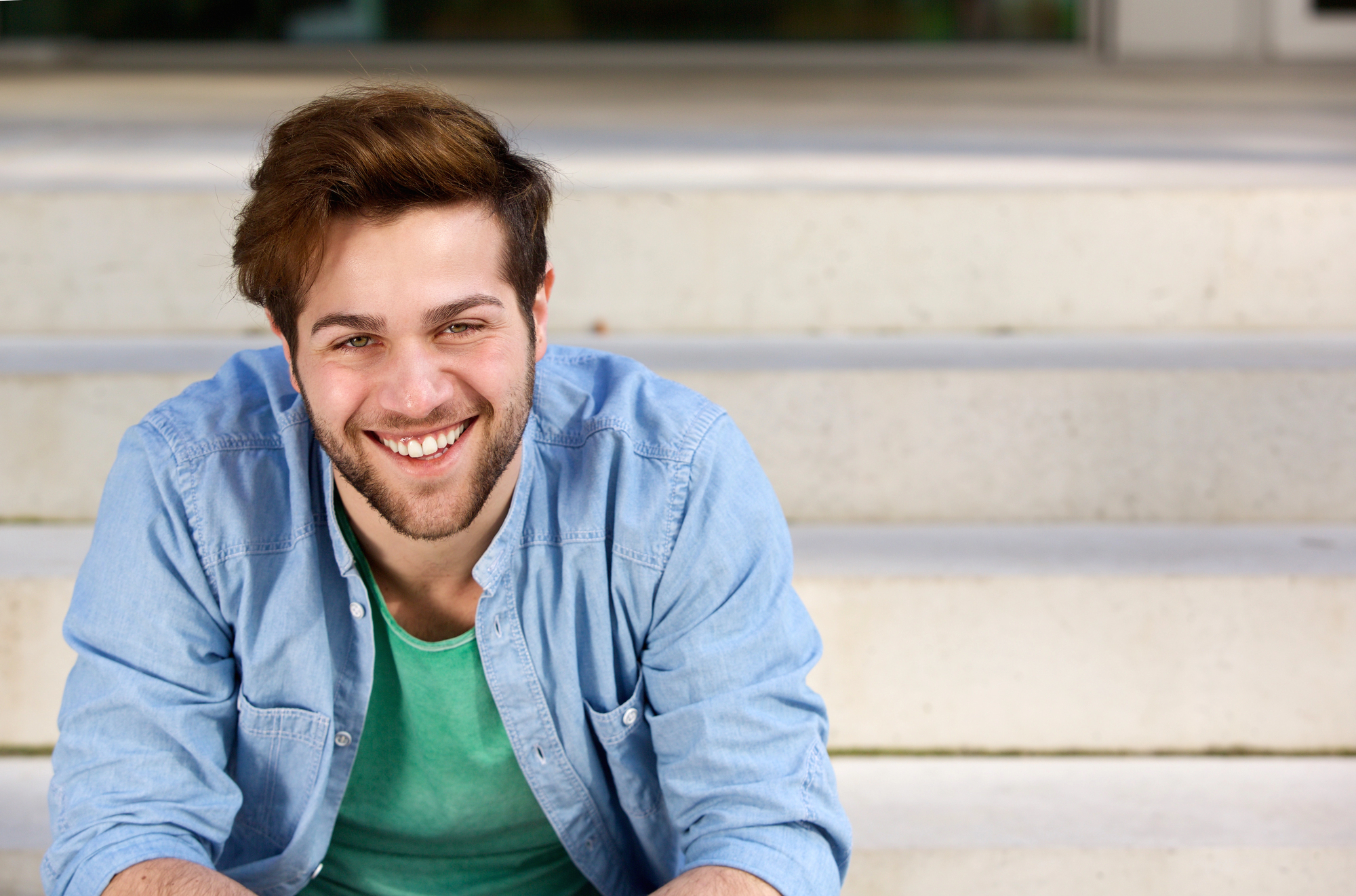 A smiling man with a beard and short, brown hair is sitting on a set of light-colored concrete steps. He is wearing a light blue, button-up shirt over a green t-shirt. The background is slightly blurred.