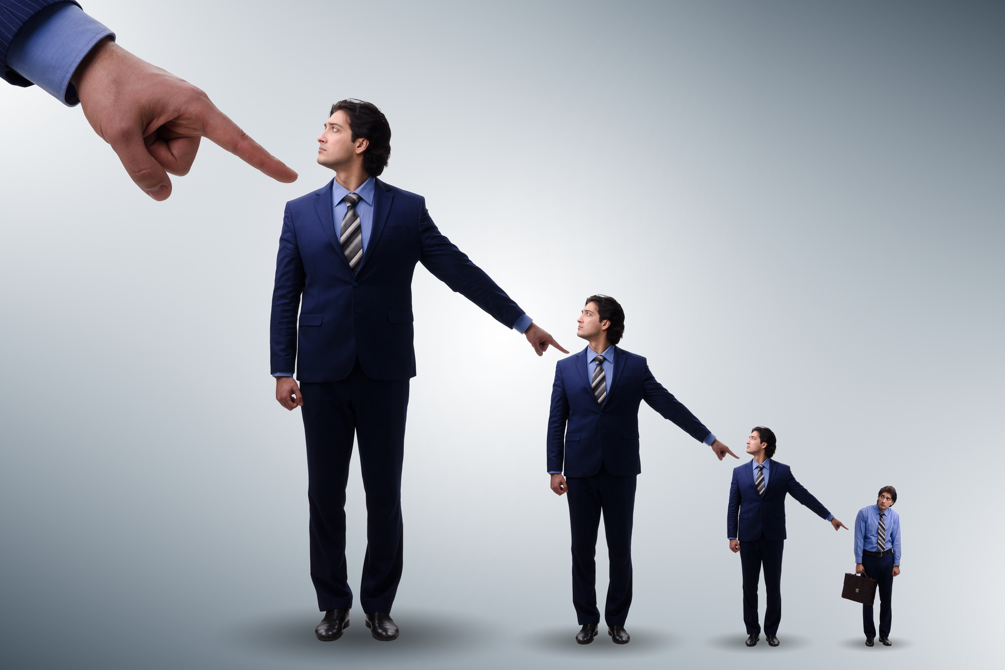 A series of four identical men in blue suits and ties are shown in a vertical line, gradually decreasing in size from left to right. The largest, on the left, is being pointed at by a large hand from above. Each man points to the smaller figure next to him.