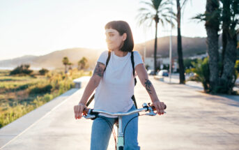 A woman with short dark hair rides a bicycle along a palm tree-lined path at sunset. She is wearing a white t-shirt and light blue jeans, carrying a backpack and has visible tattoos on her arms. The background features mountains and greenery illuminated by the setting sun.