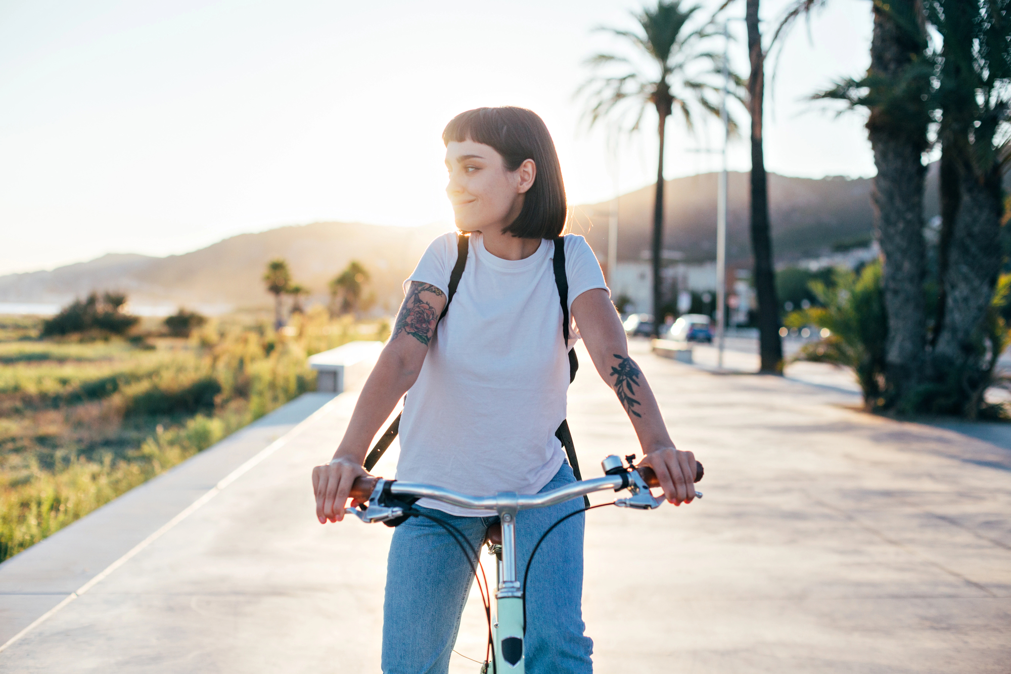 A woman with short dark hair rides a bicycle along a palm tree-lined path at sunset. She is wearing a white t-shirt and light blue jeans, carrying a backpack and has visible tattoos on her arms. The background features mountains and greenery illuminated by the setting sun.