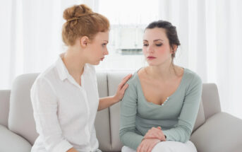 Two women sit on a couch in front of white curtains. The woman on the left, with red hair in a bun, is placing a comforting hand on the shoulder of the woman on the right, who has dark hair pulled back and is looking down with a somber expression.