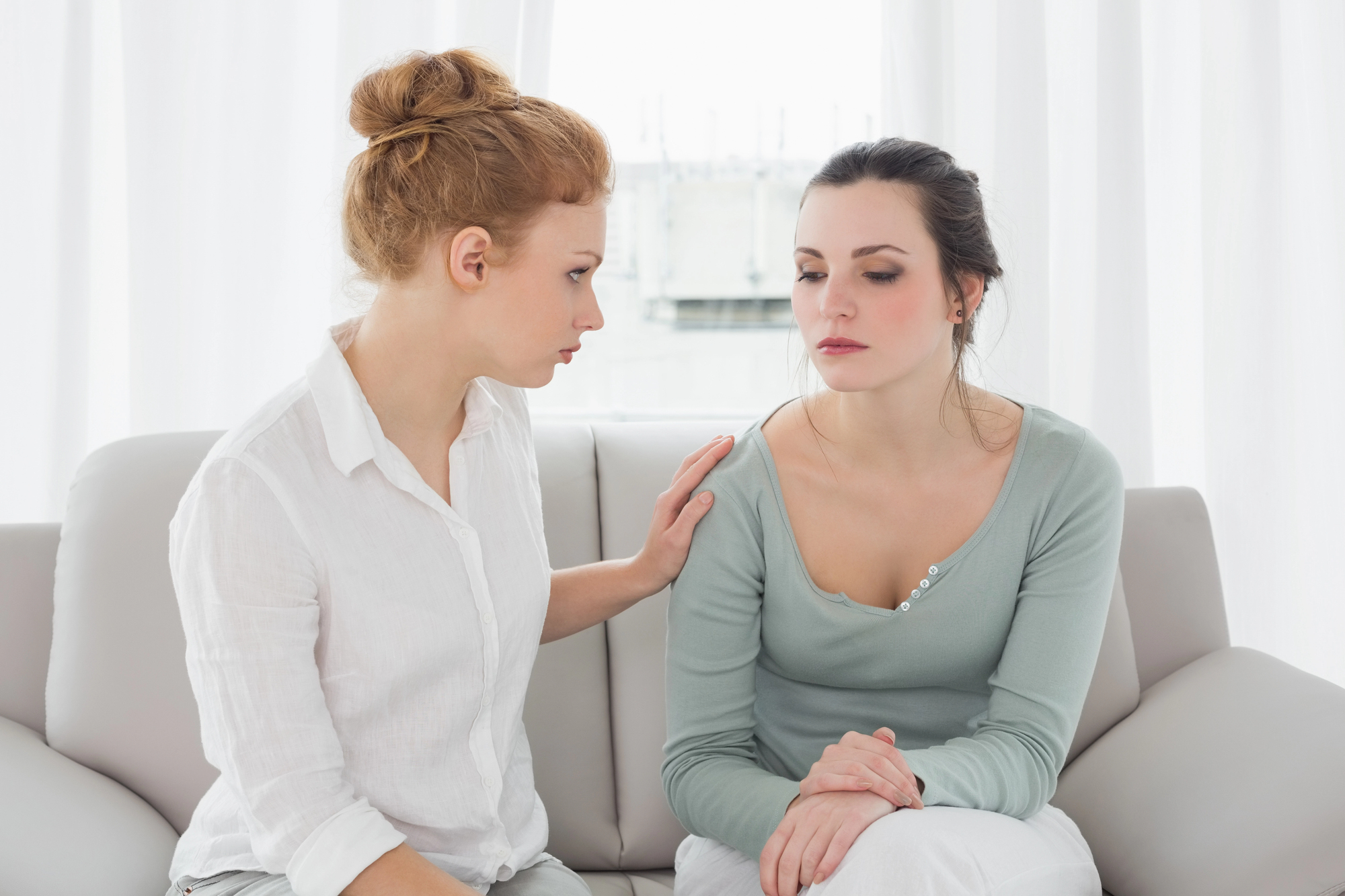 Two women sit on a couch in front of white curtains. The woman on the left, with red hair in a bun, is placing a comforting hand on the shoulder of the woman on the right, who has dark hair pulled back and is looking down with a somber expression.