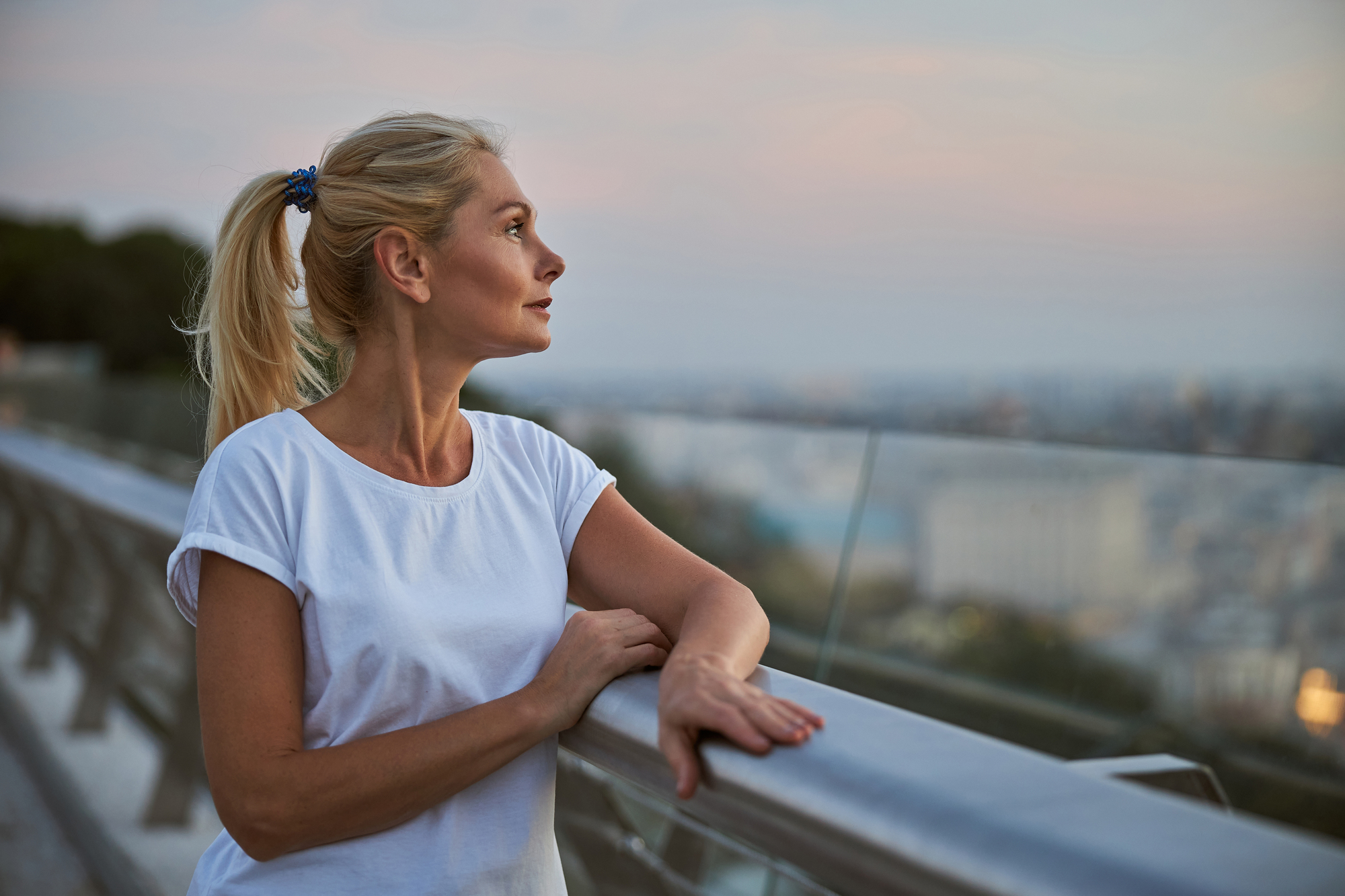 A woman with blonde hair tied in a ponytail, wearing a white t-shirt, stands by a railing and looks thoughtfully into the distance. The background features an out-of-focus cityscape under a sky at dusk.