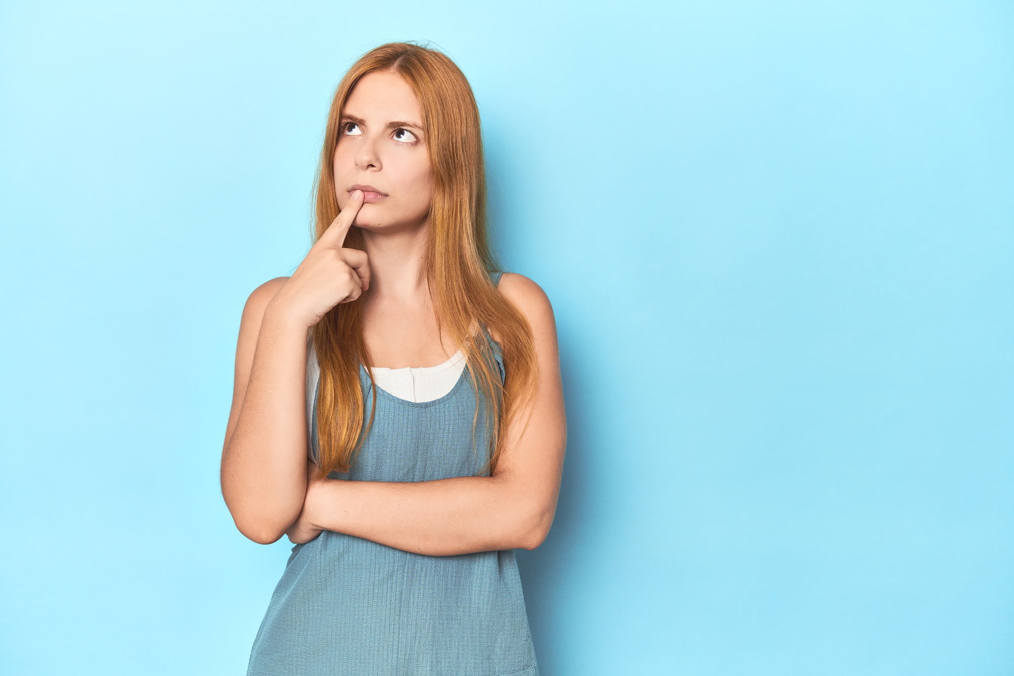 A young woman with long red hair is standing against a light blue background. She is wearing a sleeveless blue top and has one finger resting on her lips while looking upwards, appearing to be in a thoughtful or contemplative mood.