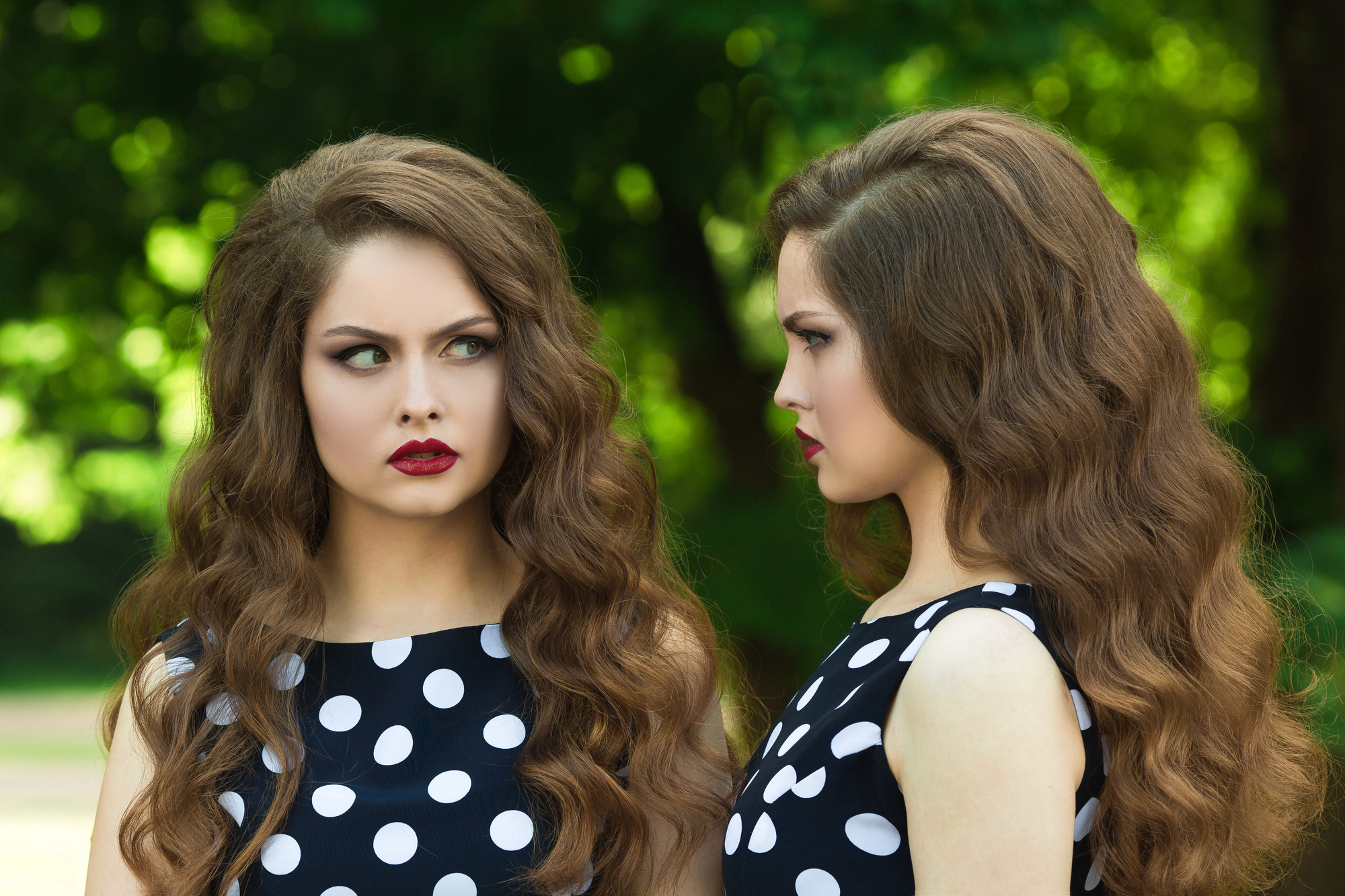 Two women with long, wavy brown hair wearing black and white polka dot dresses stand side by side. One gazes directly at the camera with a serious expression, while the other looks at her from a profile view. A lush, green outdoor background is visible.