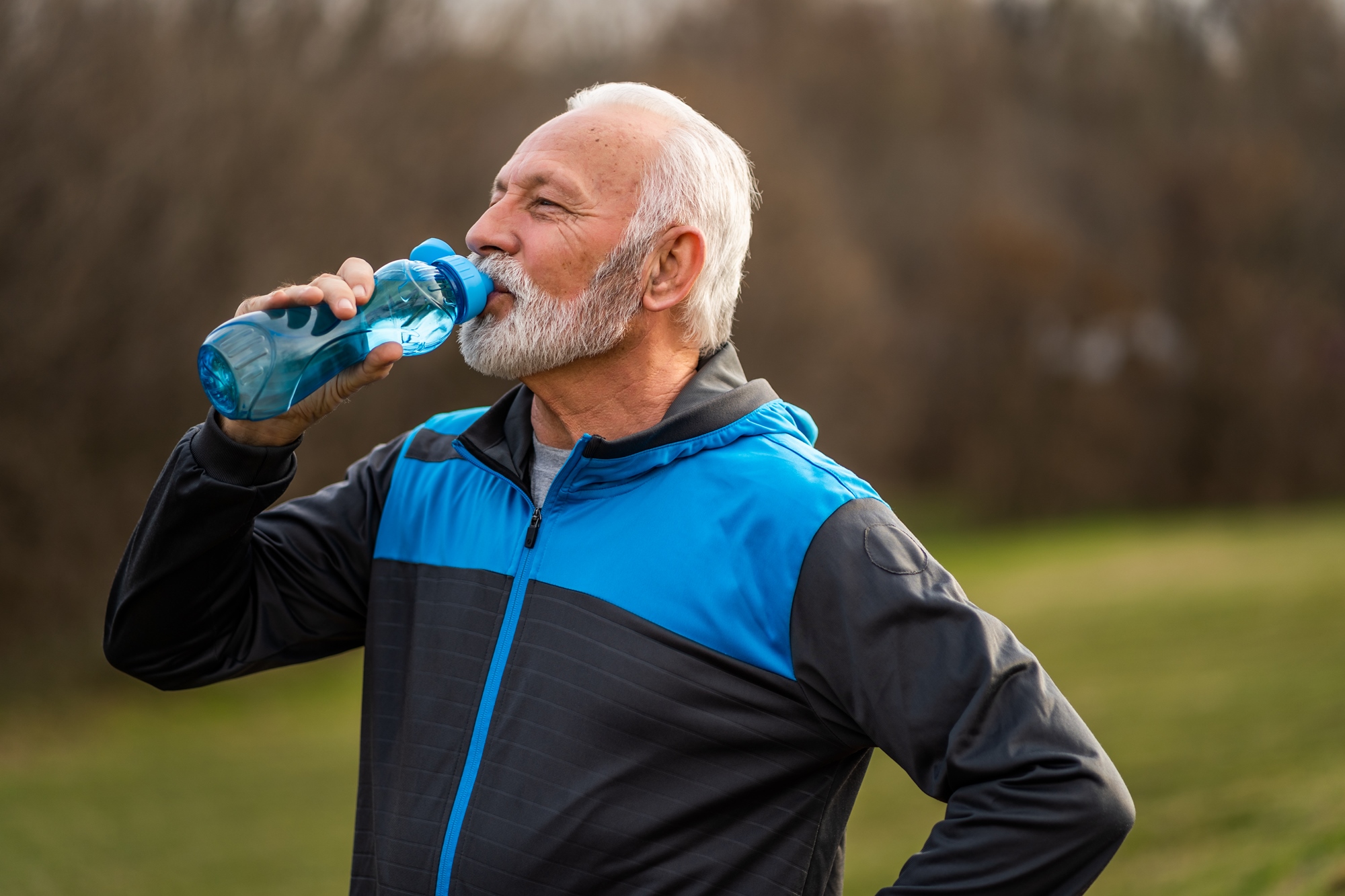 An older man with gray hair and a beard drinks from a blue water bottle. He is wearing a blue and black jacket and appears to be in an outdoor park or natural setting. His hand is on his hip, and trees are visible in the background.