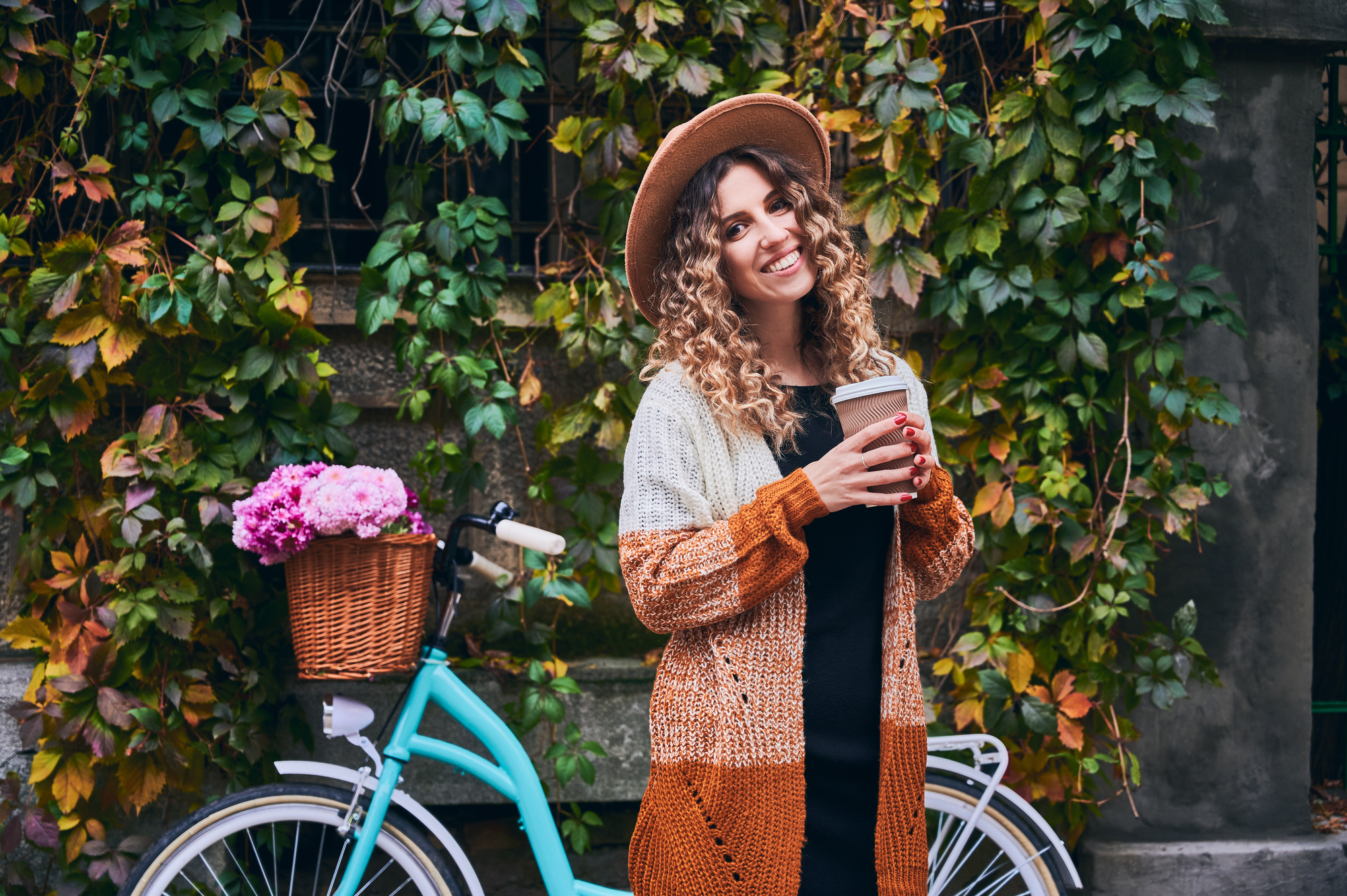 A woman with curly hair wearing a hat and a cozy, multi-colored sweater stands smiling while holding a coffee cup. Behind her is a vintage blue bicycle with a basket of pink flowers. The backdrop consists of lush green and autumnal leaves.