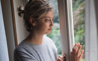 A woman with short, tied-back hair gazes thoughtfully out of a window. She is wearing a grey sweater and has her hand resting on the window frame. Green foliage is visible outside the window, suggesting it is daytime.