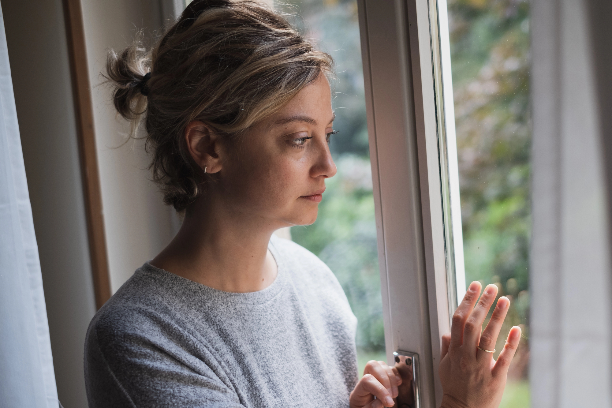 A woman with short, tied-back hair gazes thoughtfully out of a window. She is wearing a grey sweater and has her hand resting on the window frame. Green foliage is visible outside the window, suggesting it is daytime.