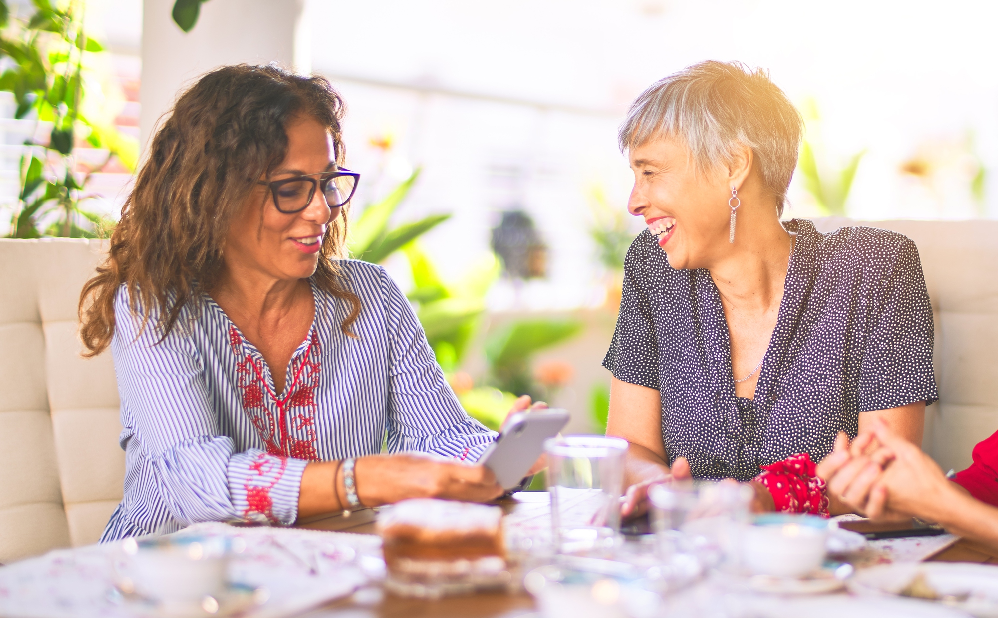 Two women sit at a table outdoors, smiling and engaging in conversation. One woman holds a smartphone, and the table is adorned with coffee cups, a cake, and other items. The background includes greenery and soft sunlight, creating a warm and cheerful atmosphere.