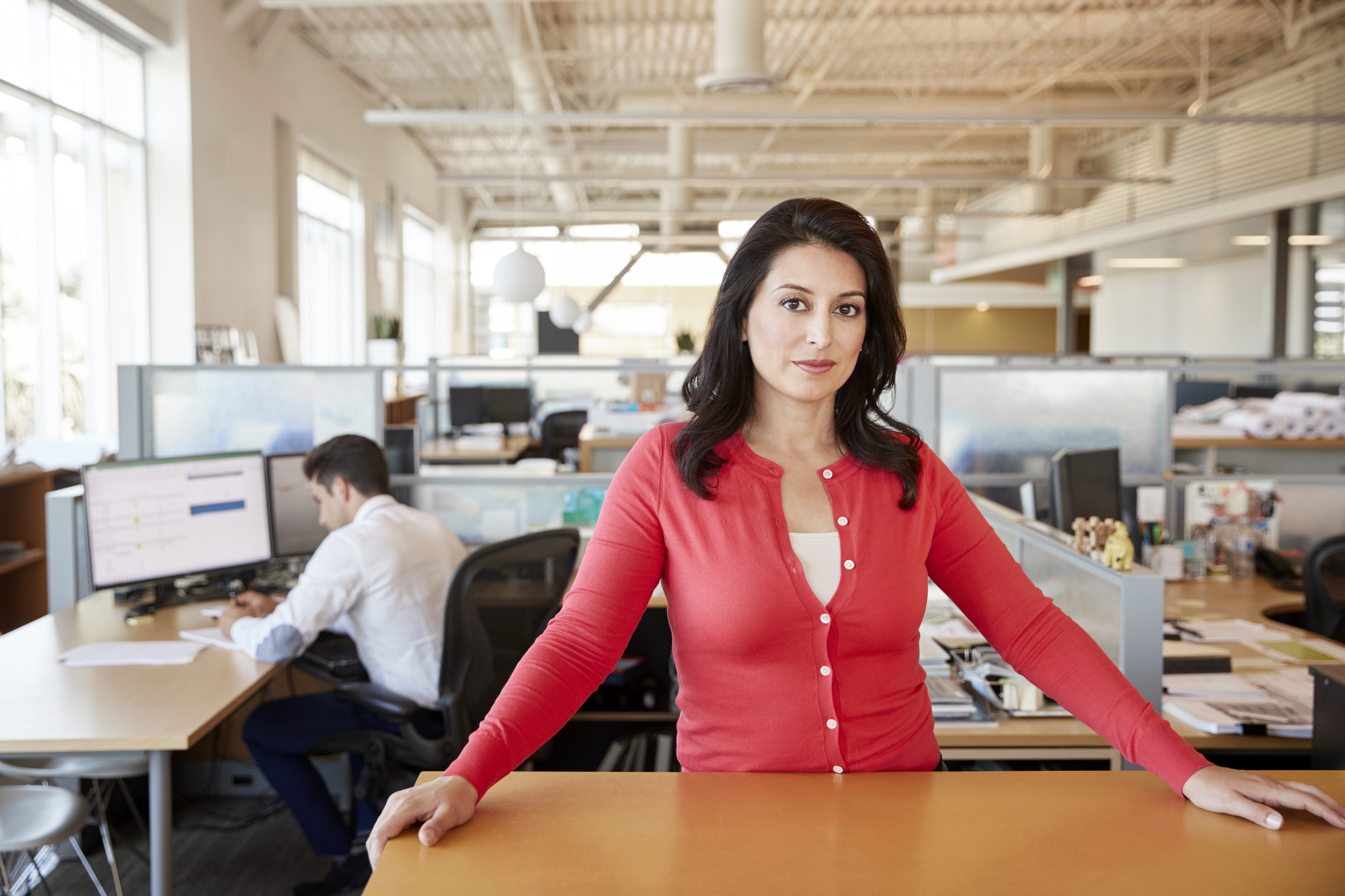 A woman in a red cardigan stands confidently, leaning on a desk in a modern, open-plan office. She looks into the camera, while in the background, a man works at a computer. The office is brightly lit with natural light from large windows.