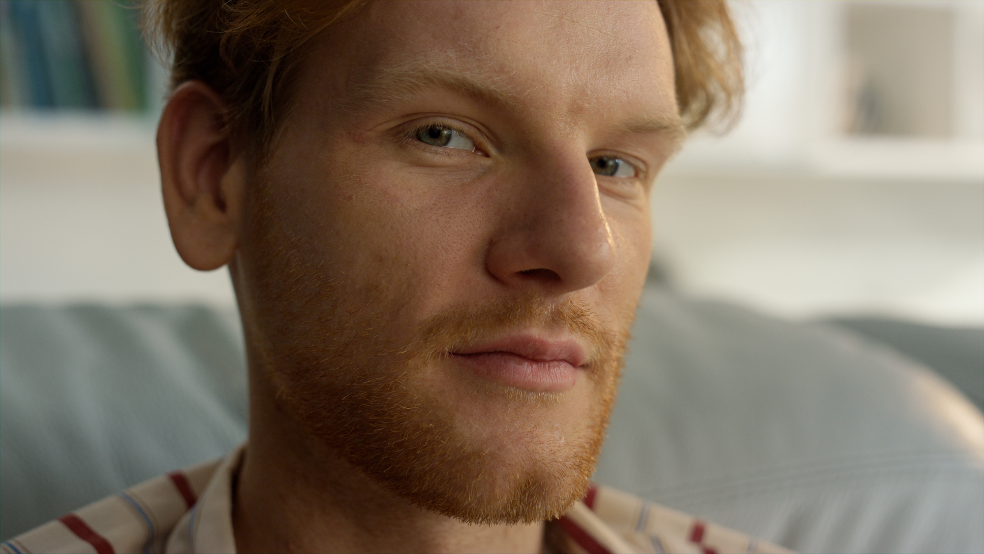 A close-up of a person with fair skin and red hair, including a red beard, looking directly at the camera with a neutral expression. The background is softly blurred, featuring an indoor setting with some indistinct light and shelves.