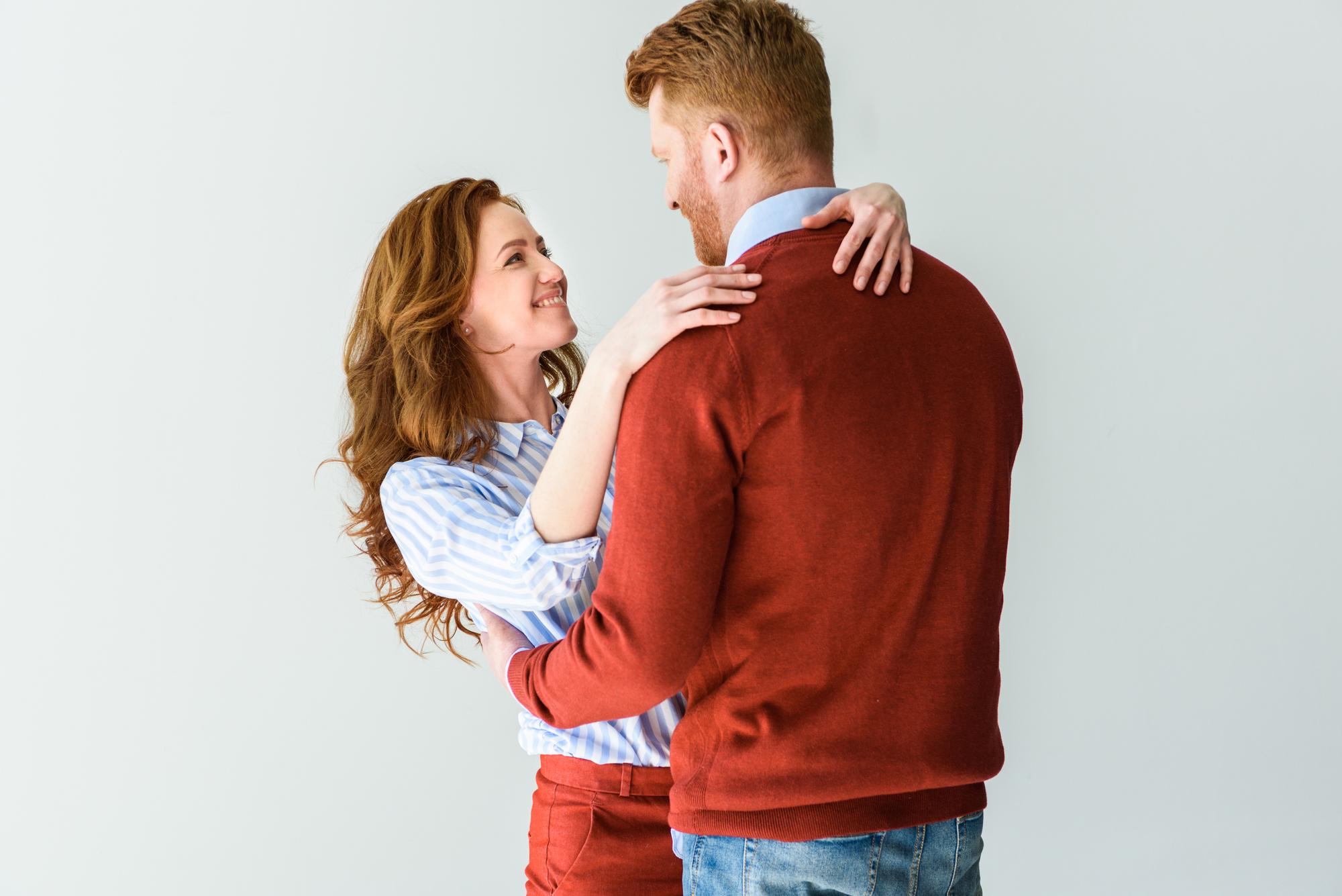 A couple stands close and embraces each other lovingly. The woman, with long wavy hair, smiles up at the man and has her arms around his shoulders. Both are wearing casual clothes; she is in a striped shirt and he is in a red sweater. They appear happy and relaxed.