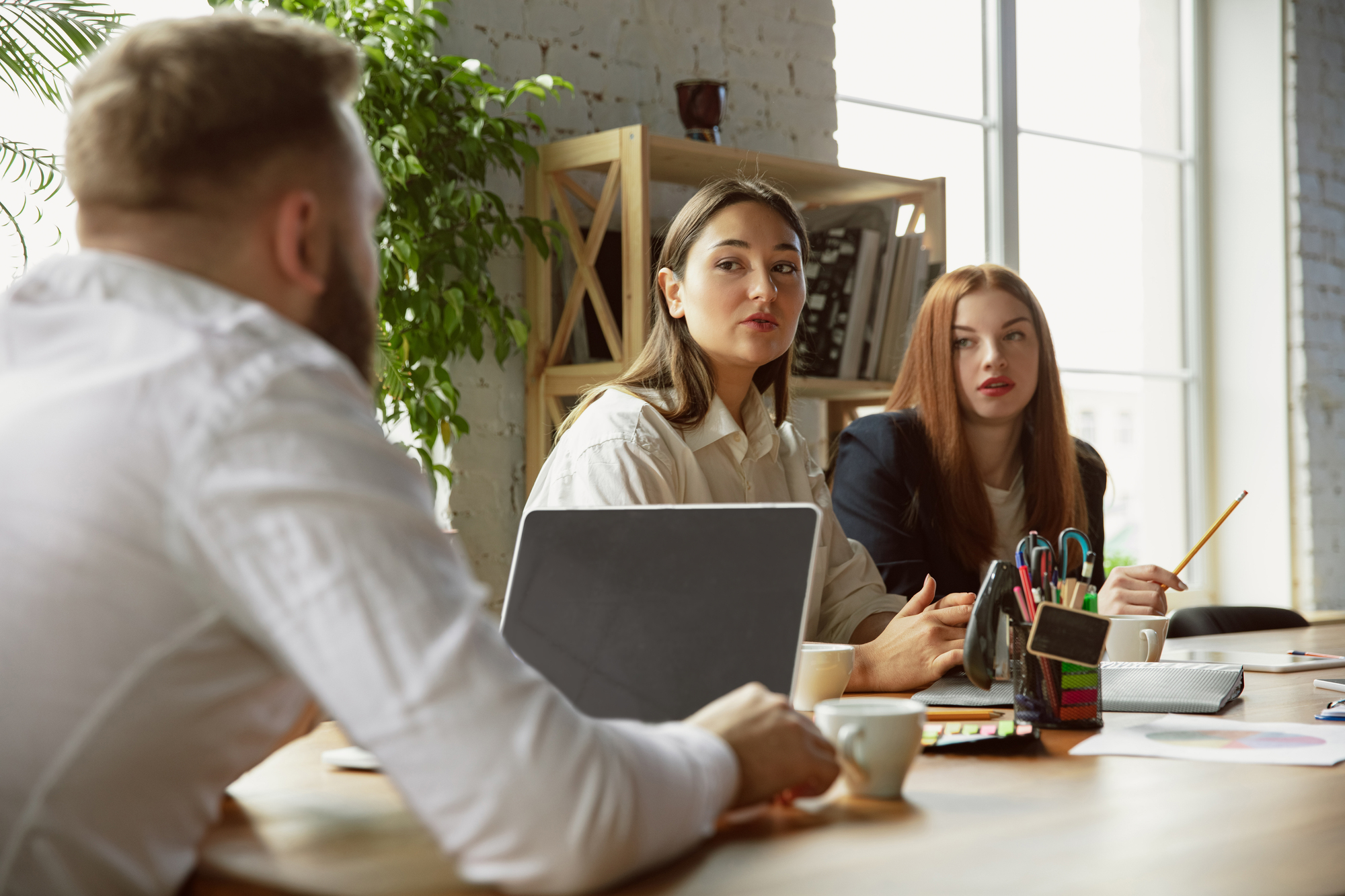 Three people are gathered around a table in a bright office space, engaging in a discussion. One man in a white shirt is seen from the back, while two women, one with brown hair and one with red hair, listen attentively. Various office supplies are on the table.