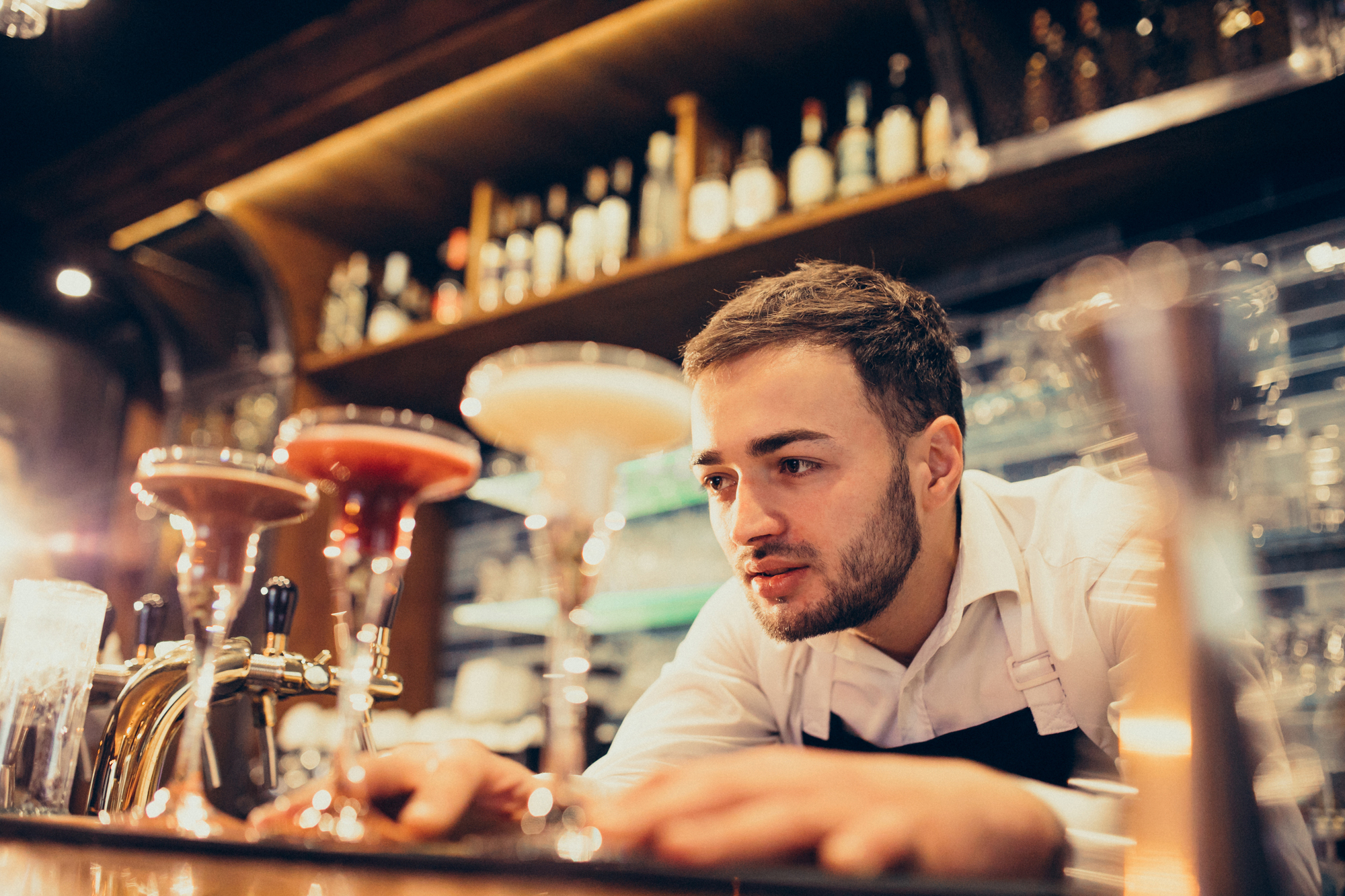 A bartender in a white shirt with rolled-up sleeves and a black apron is focused on preparing cocktails. He is standing behind a well-stocked bar with various bottles and glassware. Three colorful cocktails in large glasses are on the countertop in front of him.