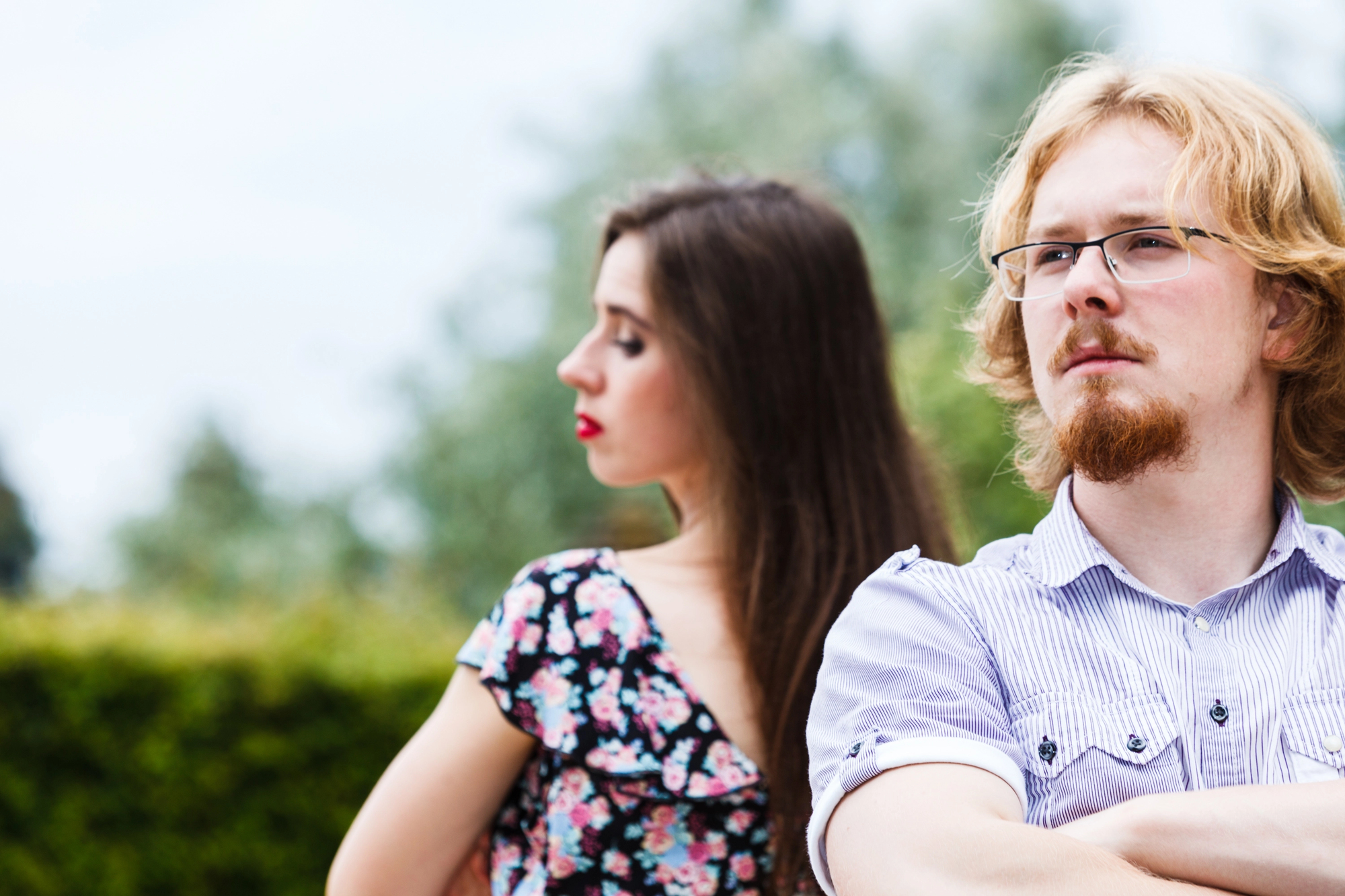 A man and a woman stand back-to-back outdoors. The man, with blond hair, a beard, glasses, and wearing a striped shirt, looks into the distance with a serious expression. The woman, with long brown hair and a floral dress, faces away, looking downward.