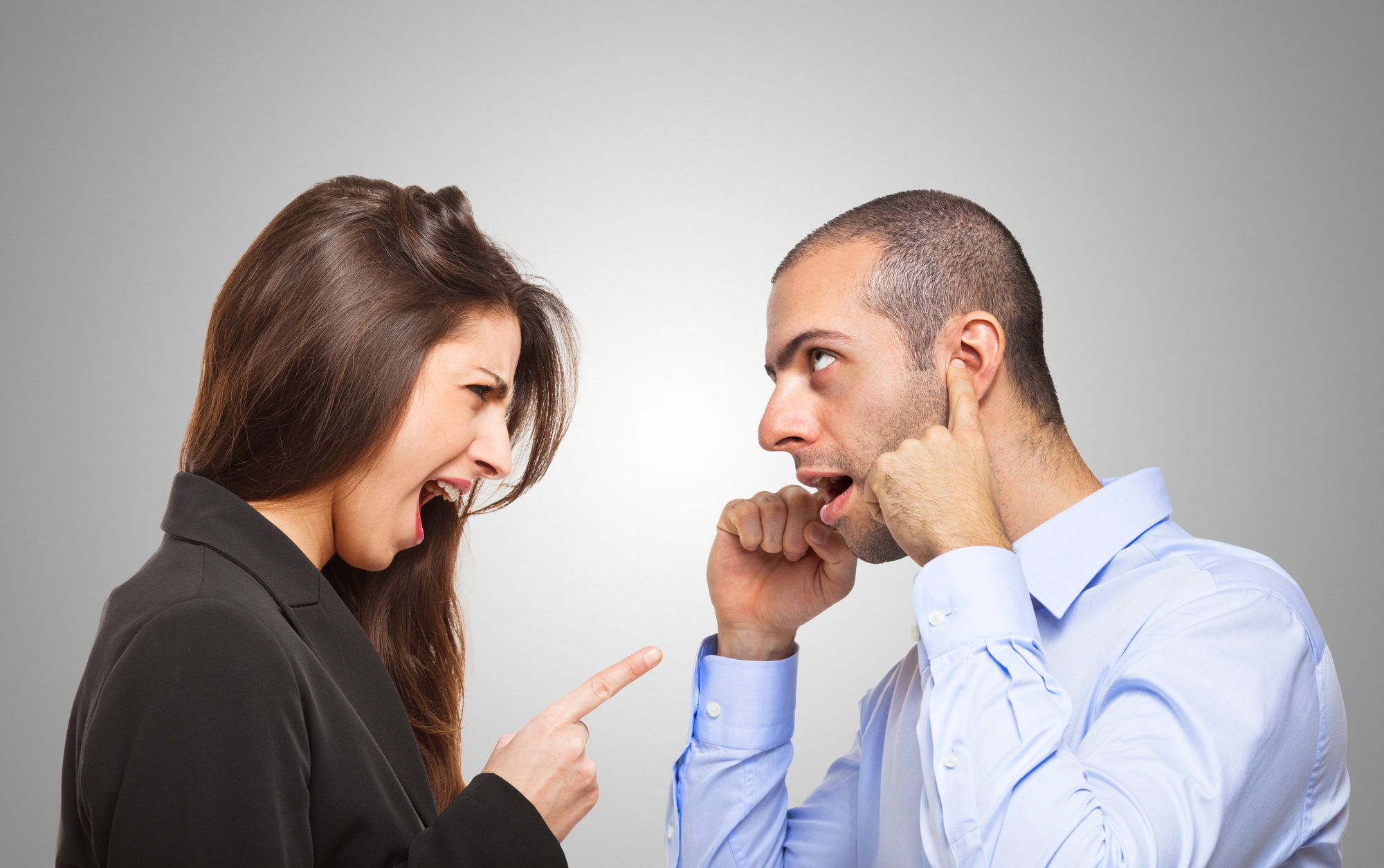 A woman with long brown hair in a black blazer is angrily pointing and yelling at a man with short hair in a light blue shirt. The man is looking surprised and plugging his ears with his fingers. Both are against a gray background.