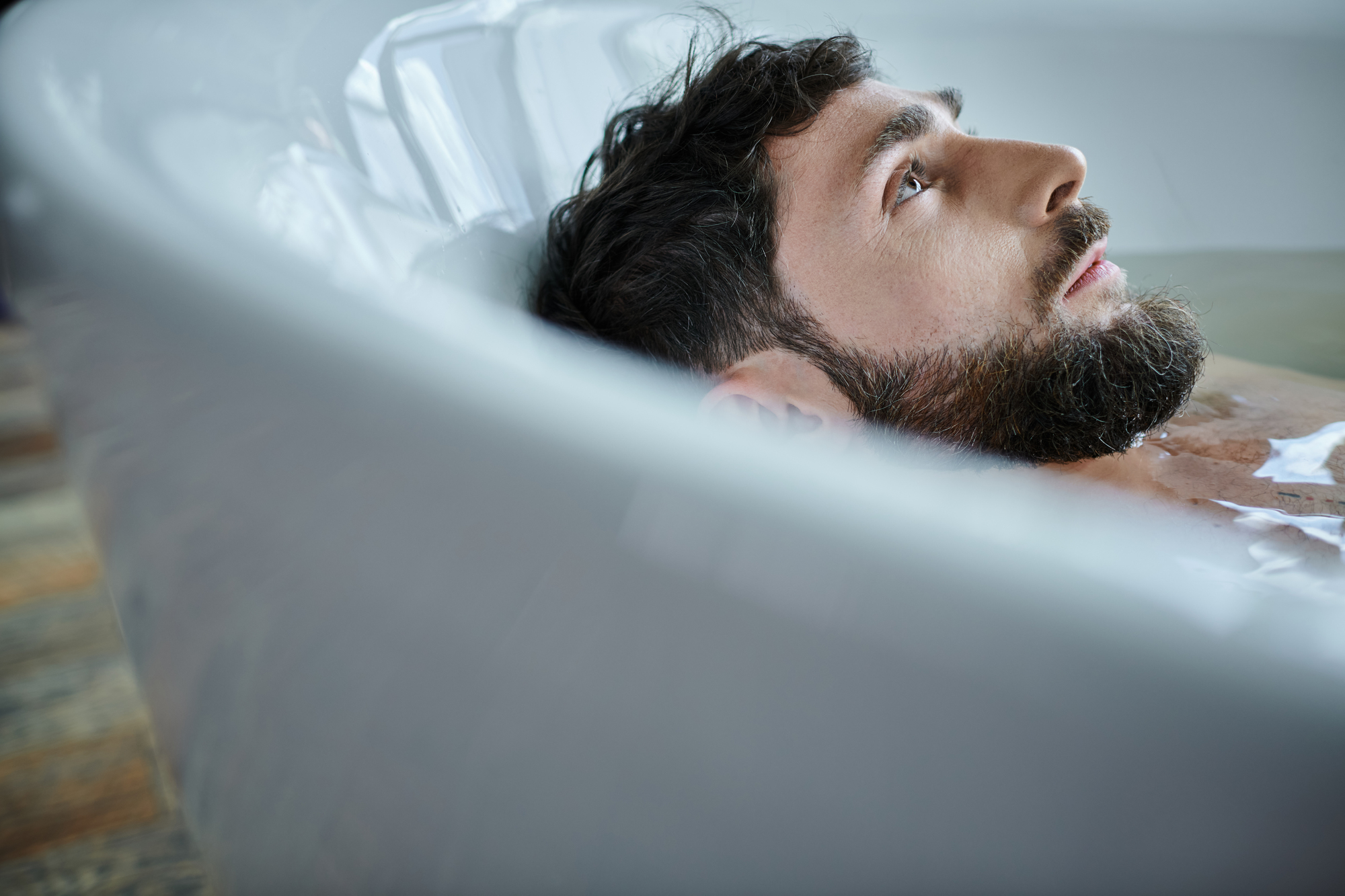 A bearded man with dark hair lies in a bathtub filled with water, looking thoughtfully upwards. Only his head and shoulders are visible above the water. The bathtub is white, and the man appears relaxed and contemplative.