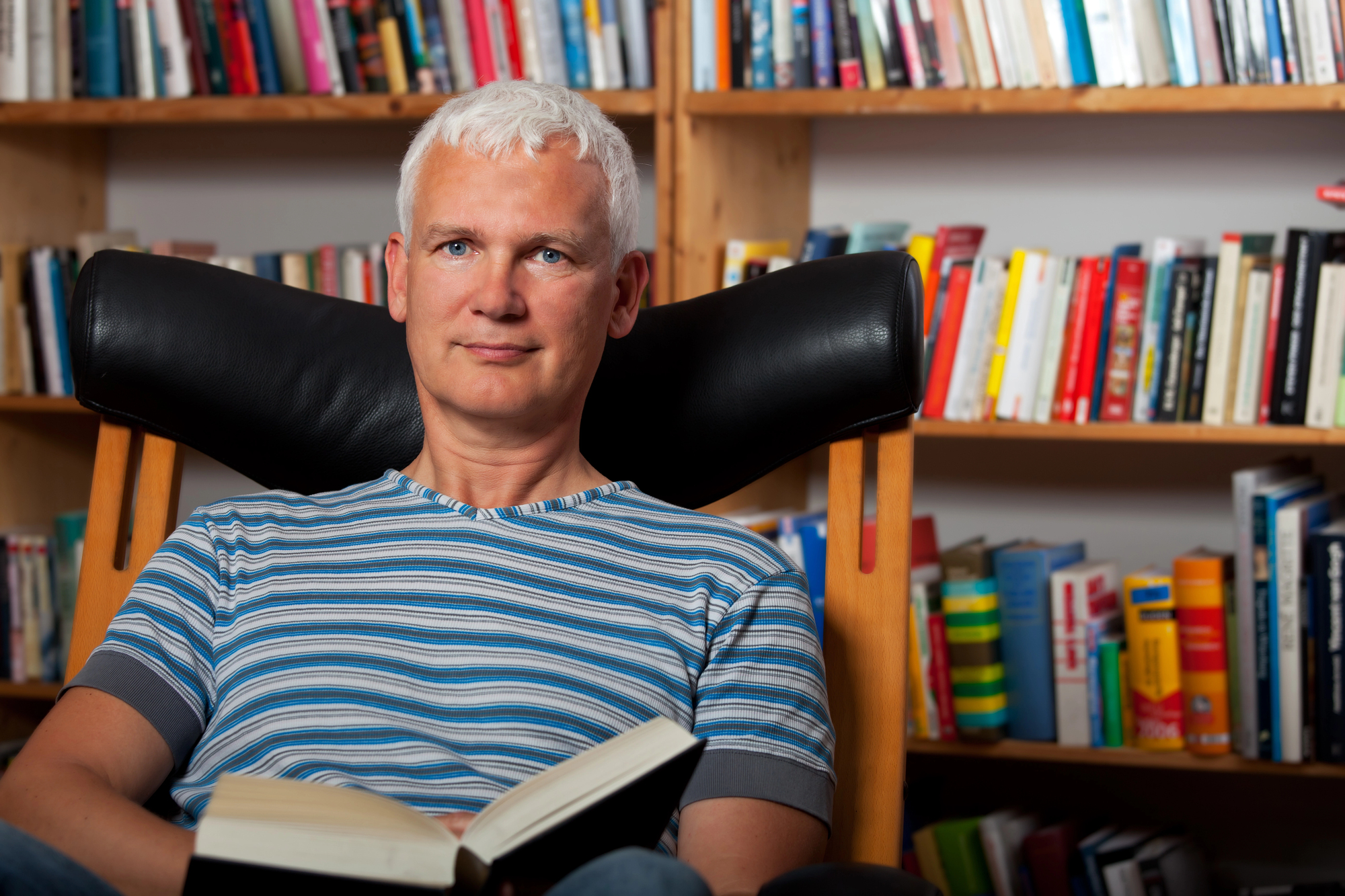 A person with short white hair sits comfortably in a black chair holding an open book. The background features a bookshelf filled with various colorful books. The person is wearing a striped shirt and appears relaxed and content.