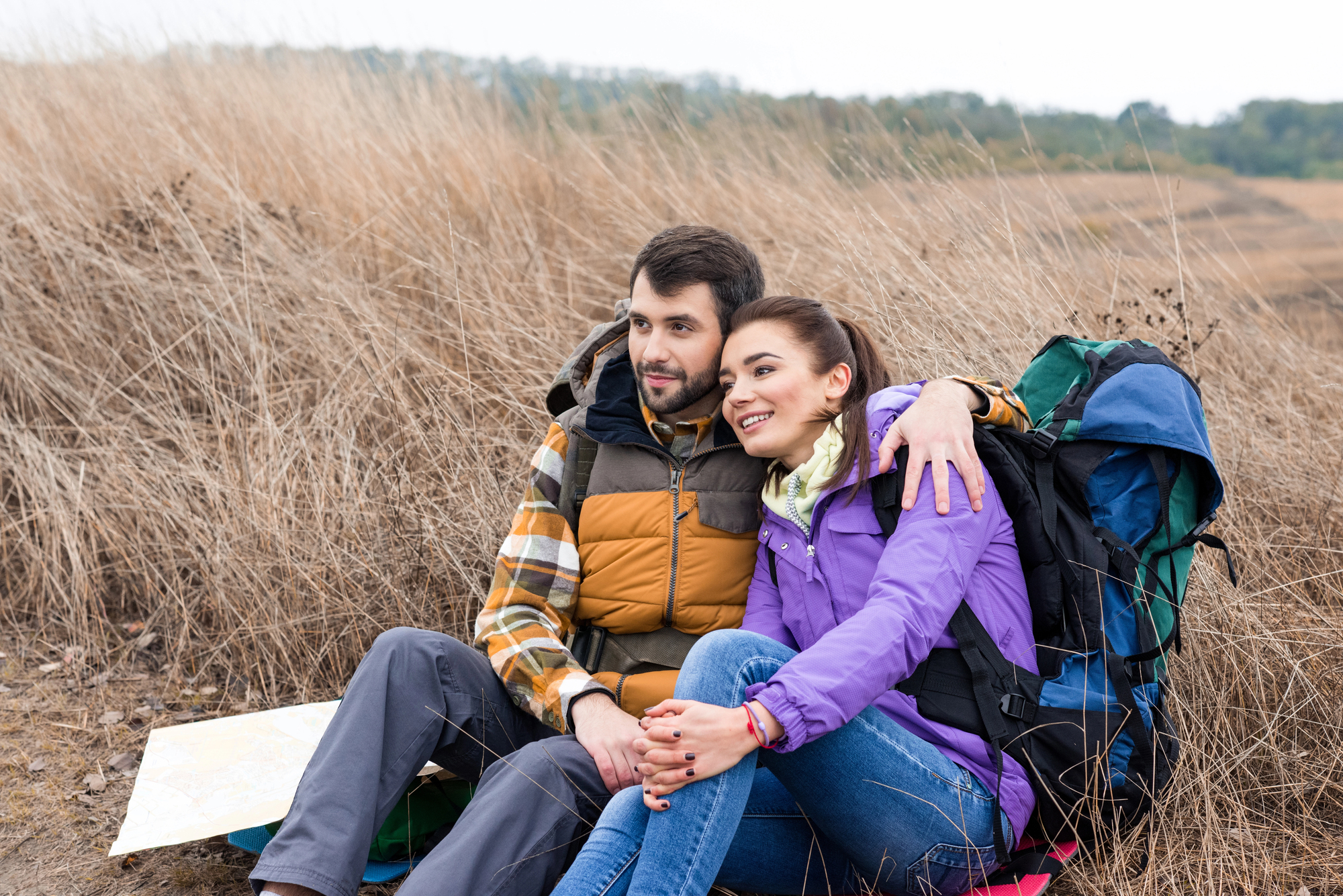 A man and a woman sit together on grassy terrain, leaning against a hill. The man wears a yellow and brown checkered jacket, and the woman wears a purple jacket. They both have hiking backpacks and look content, with the woman resting her arm around the man's shoulder.