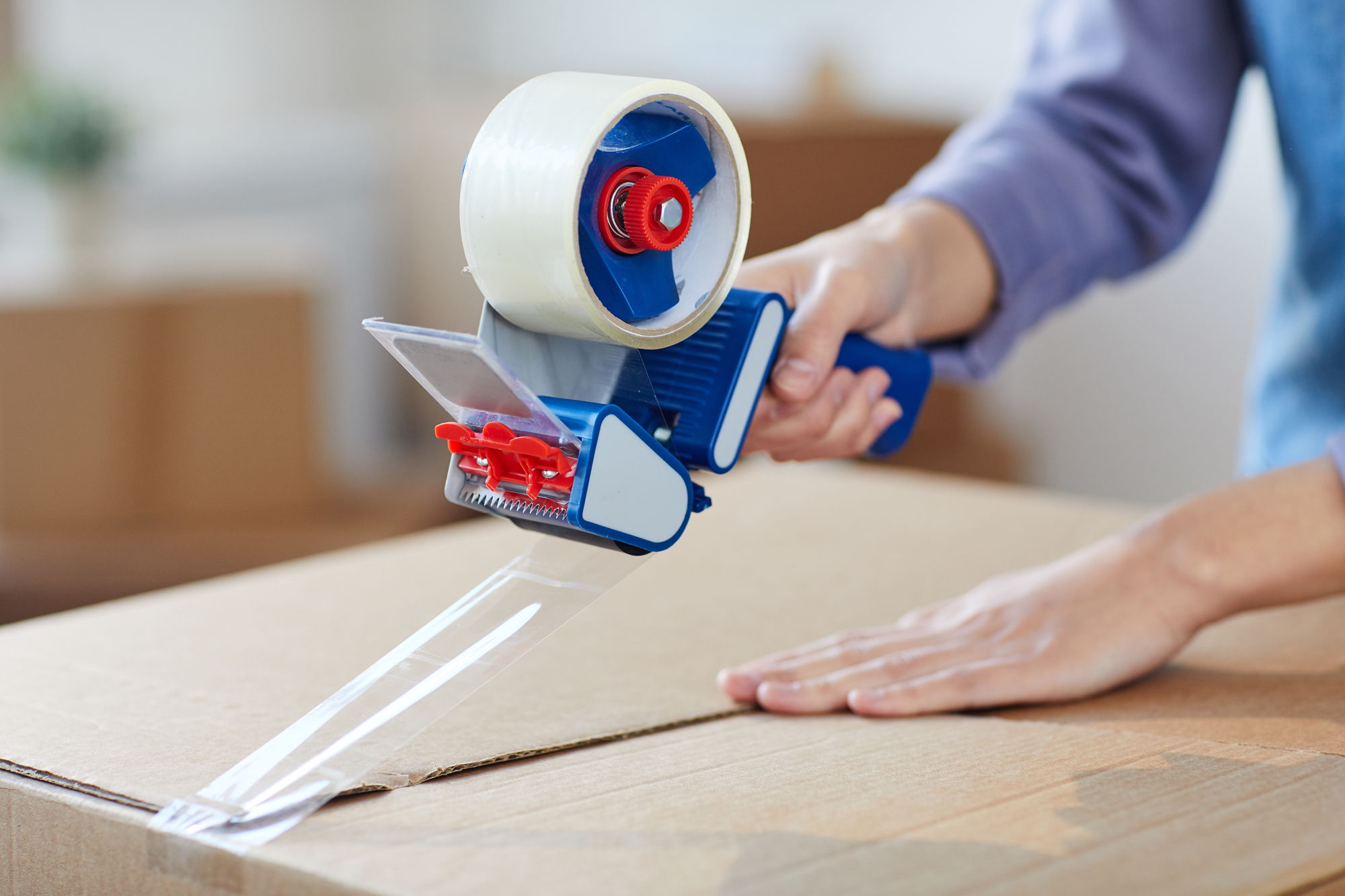 A person using a blue and red hand-held dispenser to apply clear packing tape on a cardboard box. The person’s hand smooths the tape down as it is being applied, sealing the box for shipping or storage.