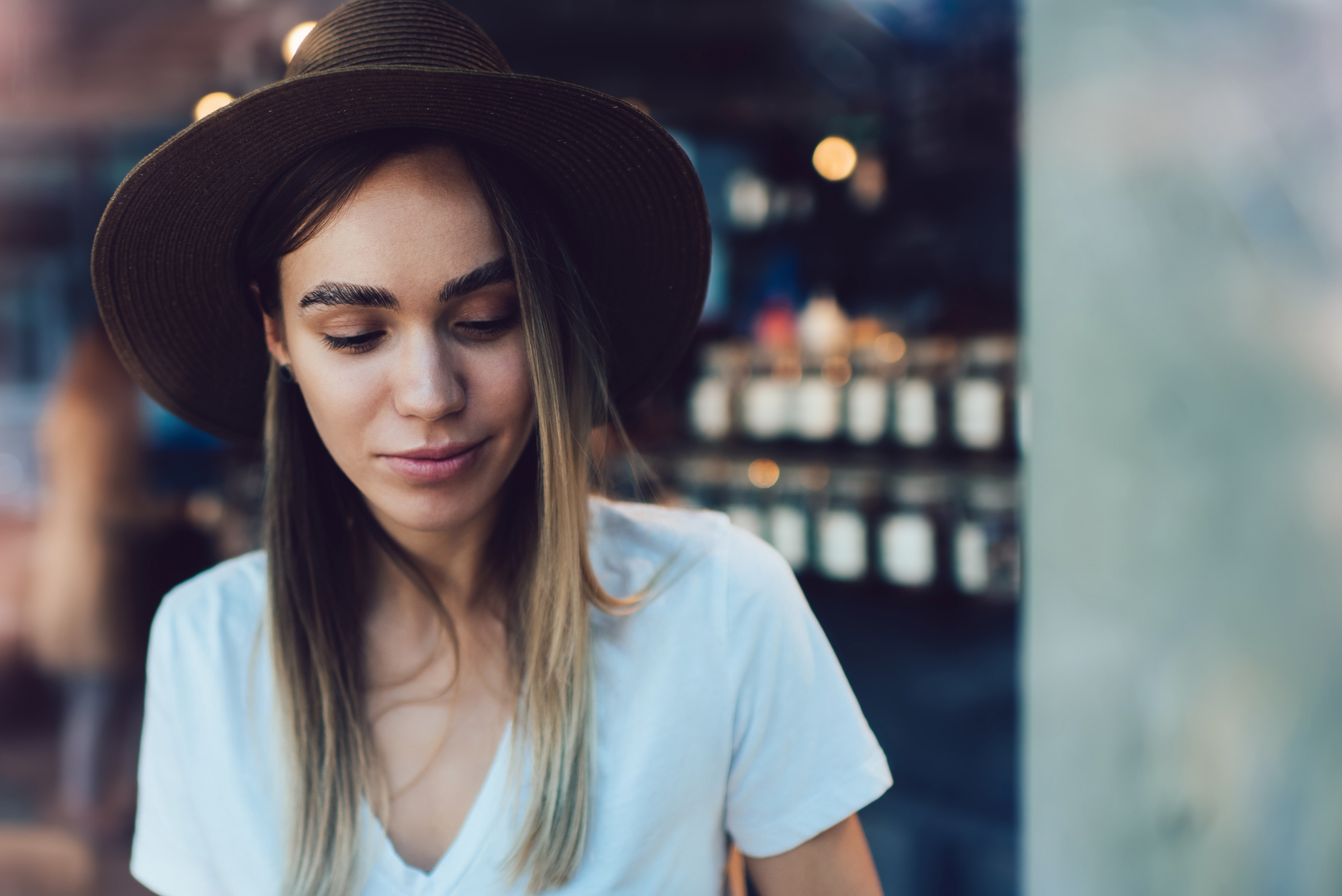 A young woman with long, straight hair wearing a brown hat and a white T-shirt is looking down while smiling slightly. The background is blurred but appears to be a shop with shelves lined with jars.