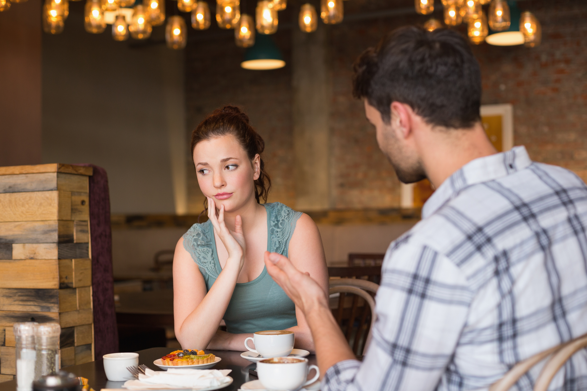 A woman and a man are sitting at a cafe table with cups of coffee and a dessert. The woman looks concerned, resting her chin on her hand, while the man is talking and gesturing with his hands. The background features warm, hanging lights and wooden decor.