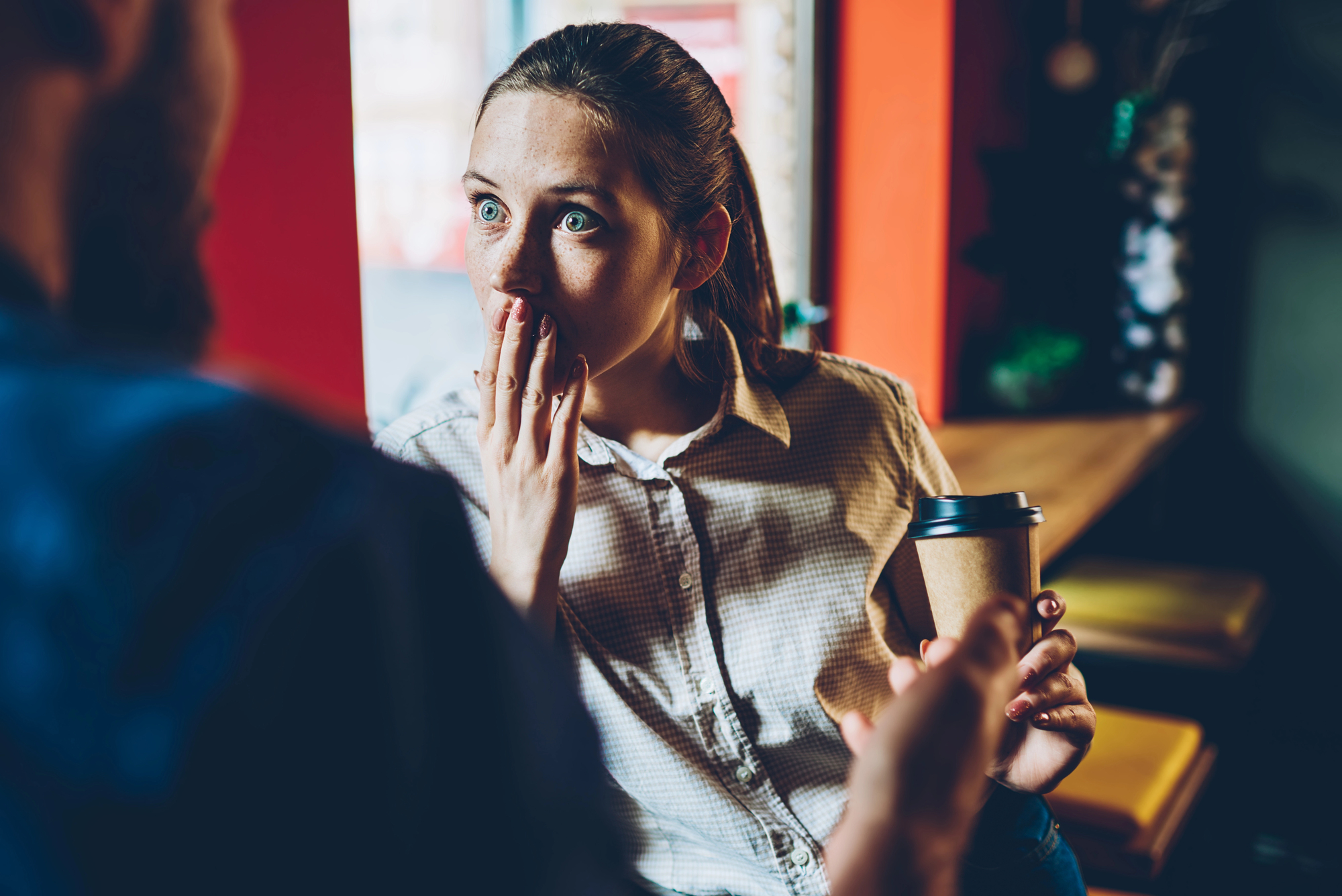 A woman in a casual outfit, holding a take-out coffee cup, is sitting indoors. She appears surprised, with her hand covering her mouth and wide eyes, as she listens to someone speaking off-screen. The background shows warm, cozy café decor.