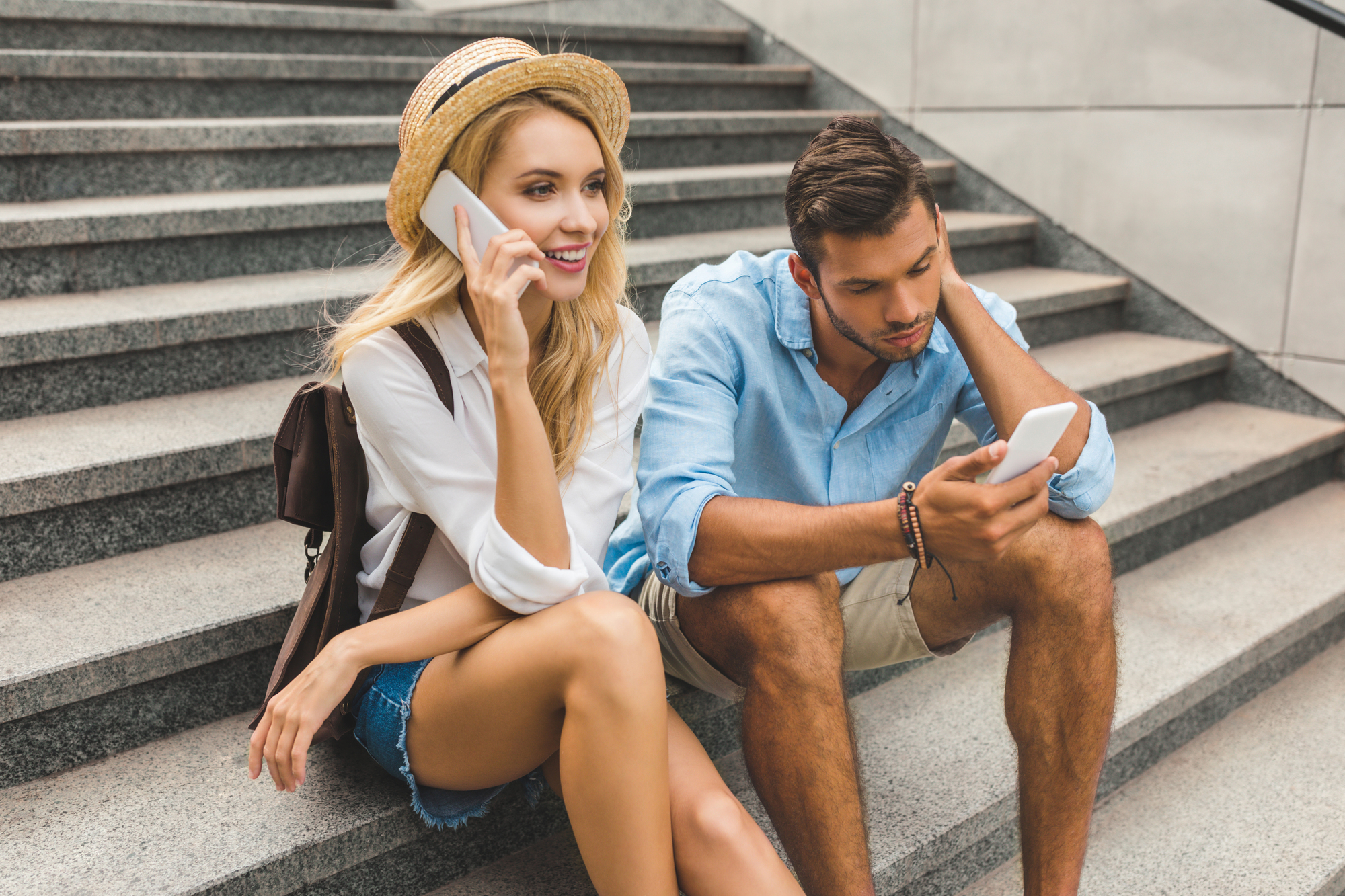 A young woman in a straw hat and denim shorts is sitting on stone steps, smiling while talking on a cellphone. Beside her, a young man in a blue shirt and shorts is sitting, looking at his cellphone with his head resting on his hand. They both have backpacks.