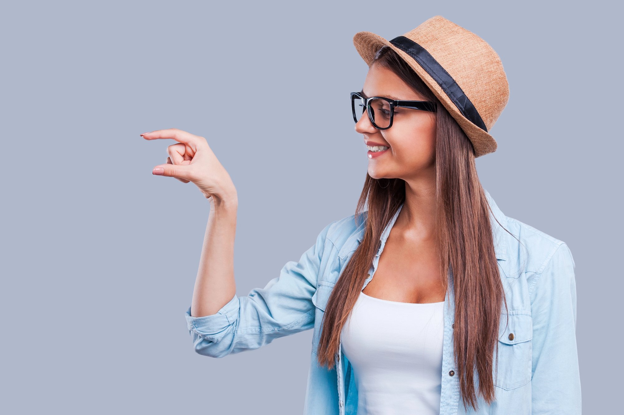 A woman wearing a straw hat, glasses, and a denim shirt over a white top is smiling and holding her fingers close together as if pinching something small. The background is a solid light grey color.