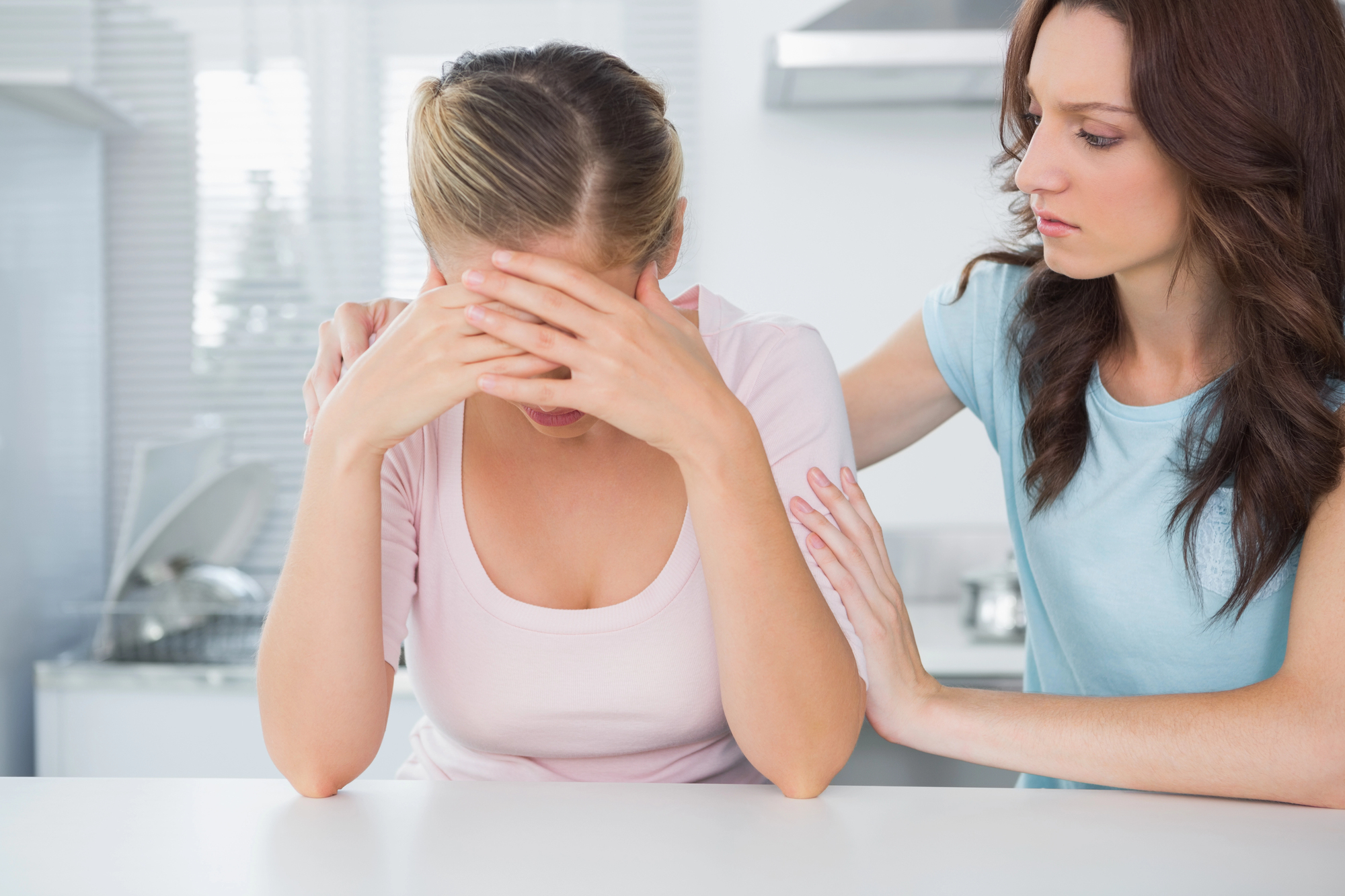 A woman with blond hair wearing a pink shirt sits at a table with her head in her hands, appearing distressed. Another woman with brown hair and a blue shirt stands beside her, offering comfort by placing a hand on her shoulder.