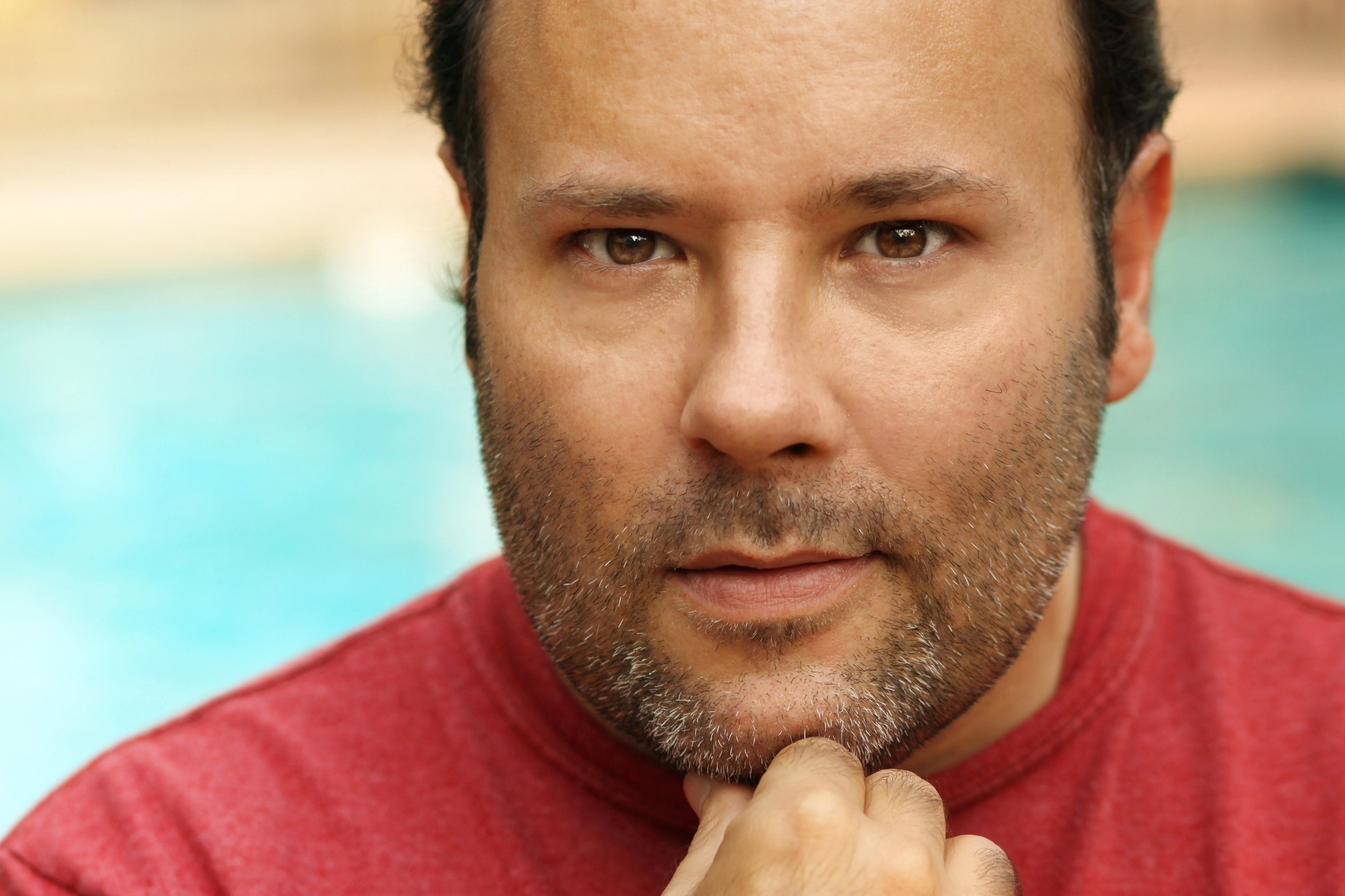 A man with short hair and a beard, wearing a red shirt, looks directly at the camera with a thoughtful expression, resting his chin on his hand. The background is blurred but suggests an outdoor setting near a body of water.