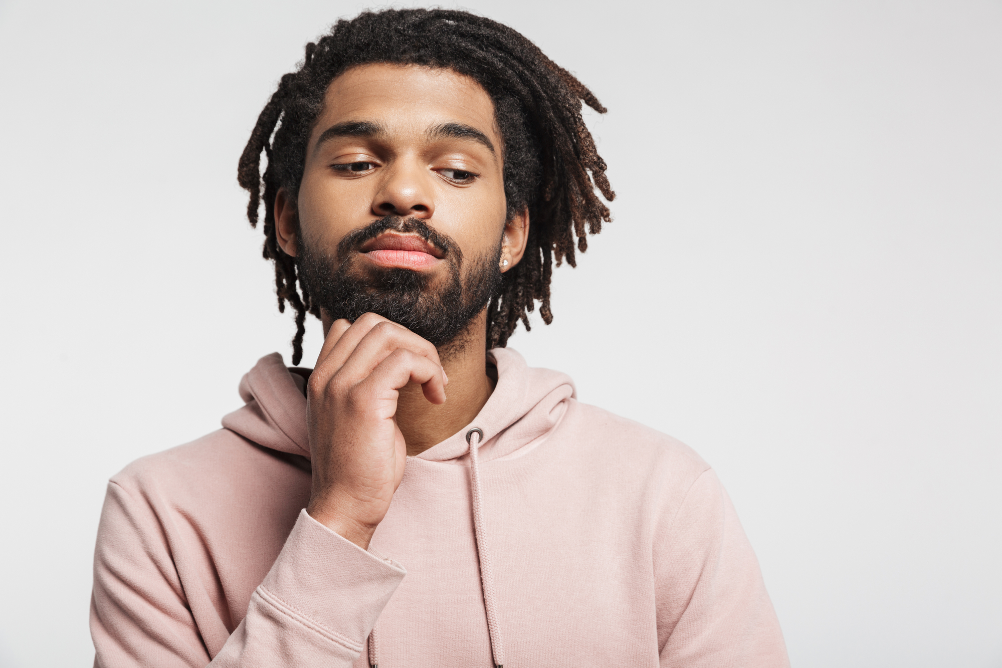 A person with dreadlocks and a beard is wearing a light pink hoodie. They are looking down and touching their chin thoughtfully against a plain white background.