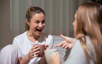 Two women sit on a couch in a cozy setting, smiling and talking animatedly. The woman on the left holds a pink mug with both hands, looking excited, while the woman on the right holds a white mug and gestures with her hand. Soft lighting creates a warm atmosphere.