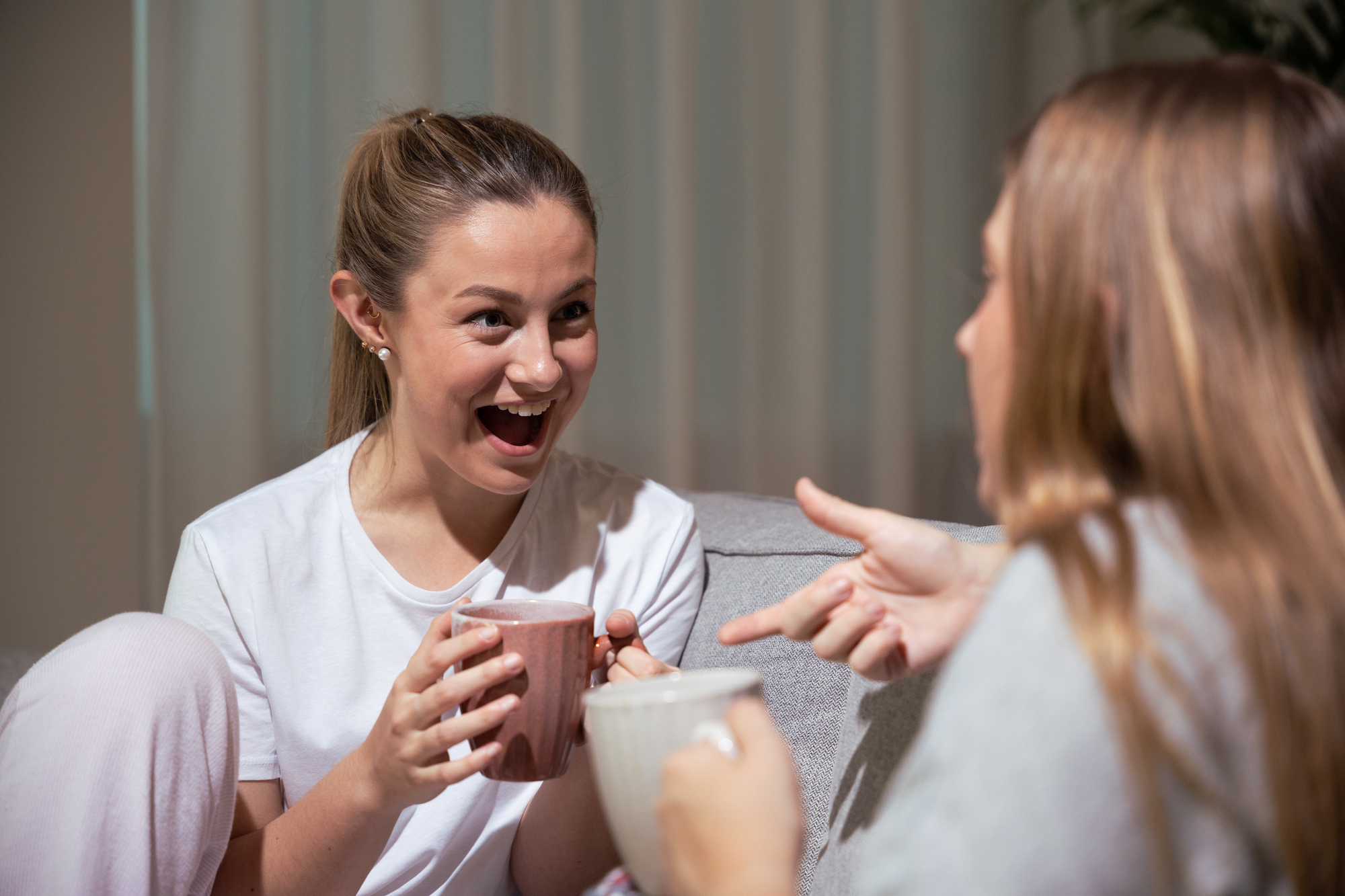 Two women sit on a couch in a cozy setting, smiling and talking animatedly. The woman on the left holds a pink mug with both hands, looking excited, while the woman on the right holds a white mug and gestures with her hand. Soft lighting creates a warm atmosphere.