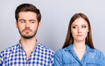 A man with dark hair and a beard wearing a blue plaid shirt and a woman with long brown hair wearing a denim shirt are standing side by side. They are looking at each other with serious expressions against a plain, light gray background.