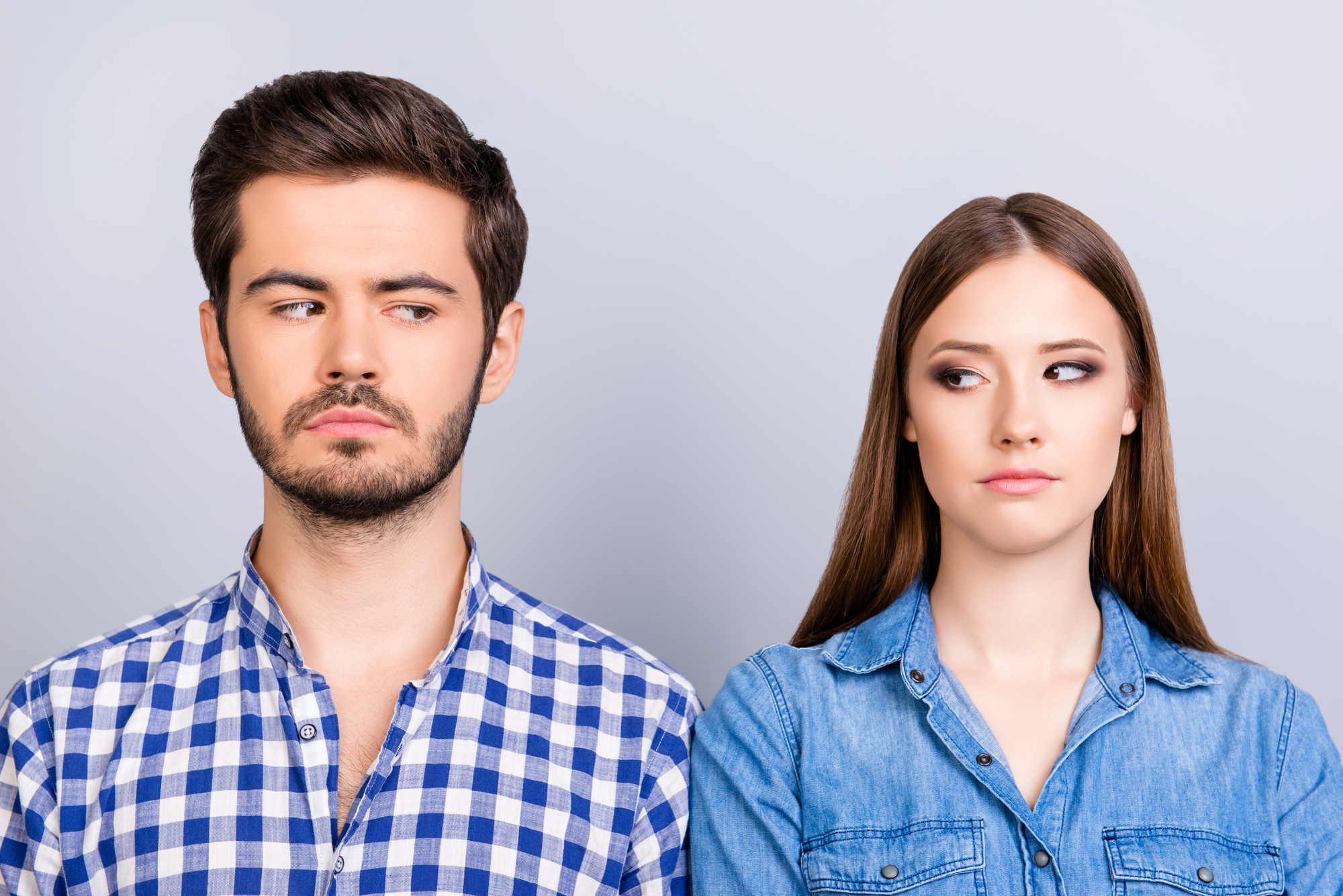 A man with dark hair and a beard wearing a blue plaid shirt and a woman with long brown hair wearing a denim shirt are standing side by side. They are looking at each other with serious expressions against a plain, light gray background.