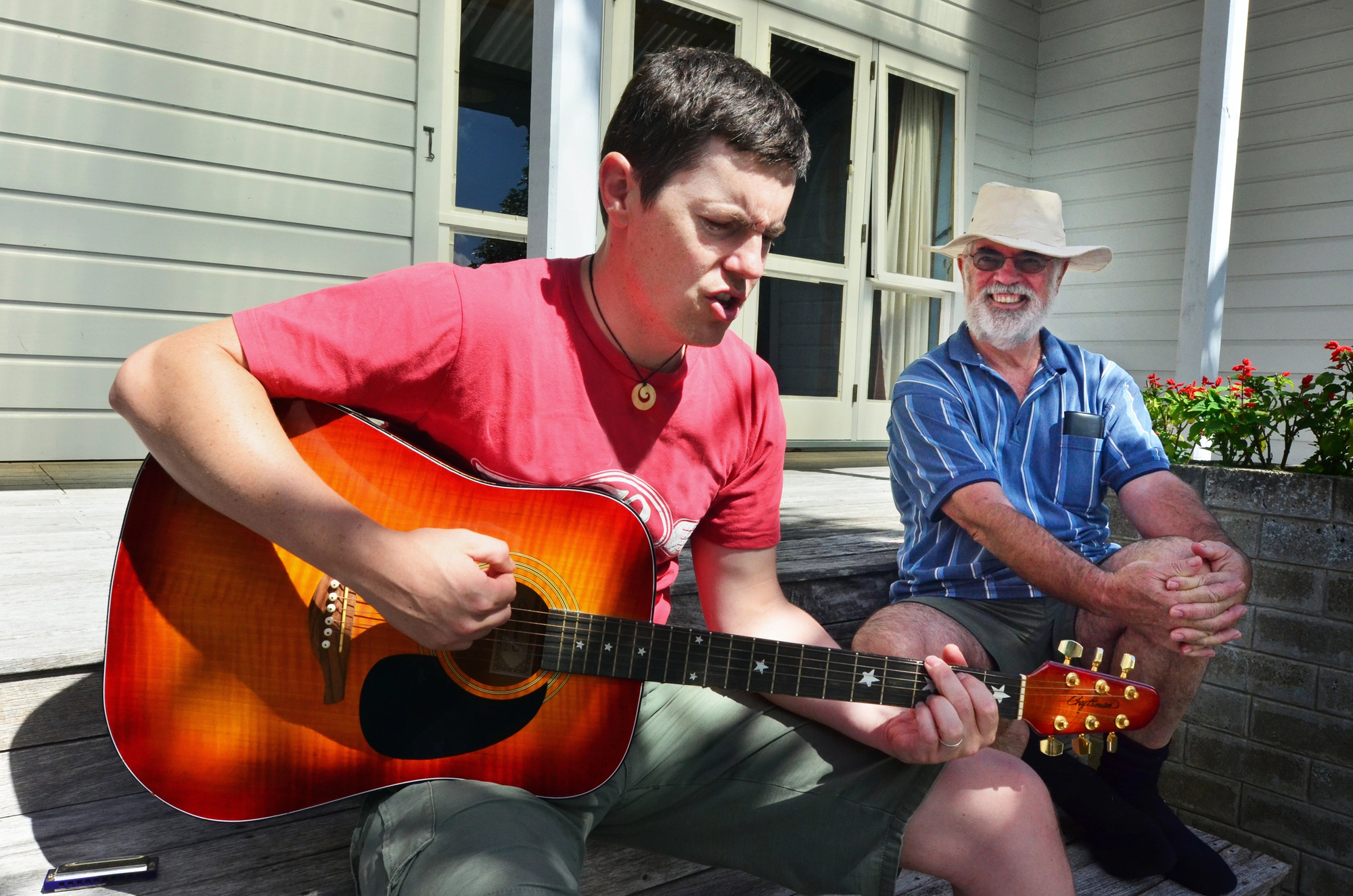 A man in a red shirt strums an acoustic guitar on a sunny porch. He is focused on playing, while an older man in a striped blue shirt and white hat sits nearby, smiling and listening. The background features the exterior of a house with flowers in bloom.