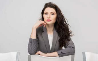 A woman in a gray blazer sits on a chair facing the camera, resting her chin on her hand, against a plain white background. She has long, dark wavy hair and a neutral expression.