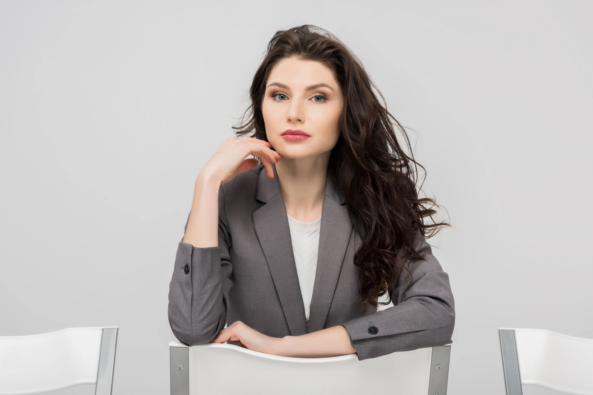 A woman in a gray blazer sits on a chair facing the camera, resting her chin on her hand, against a plain white background. She has long, dark wavy hair and a neutral expression.