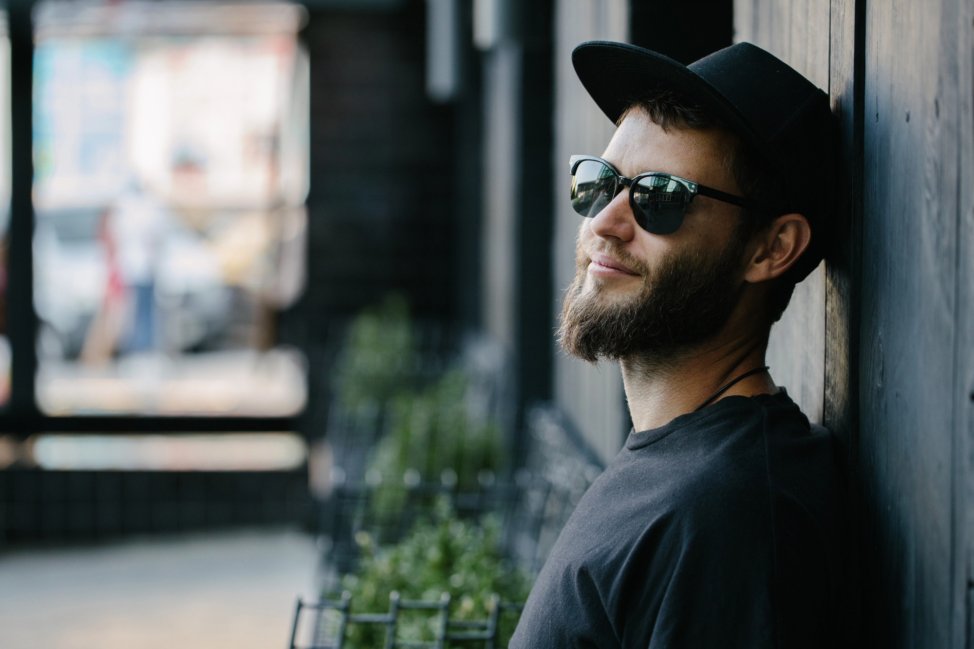 A bearded man wearing sunglasses and a black cap leans against a wooden wall. He is dressed in a black shirt and appears relaxed, with a slight smile on his face. The background is blurred but shows some outdoor elements and a row of potted plants.