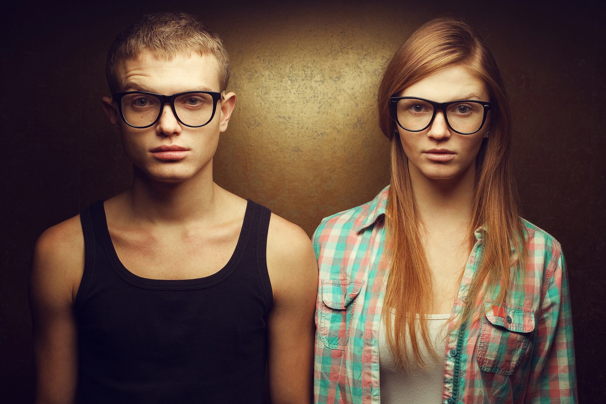 A young man and woman stand side by side against a dark, textured background. Both wear large black-rimmed glasses and have serious expressions. The man is in a black tank top, and the woman is in a checkered shirt over a white top.