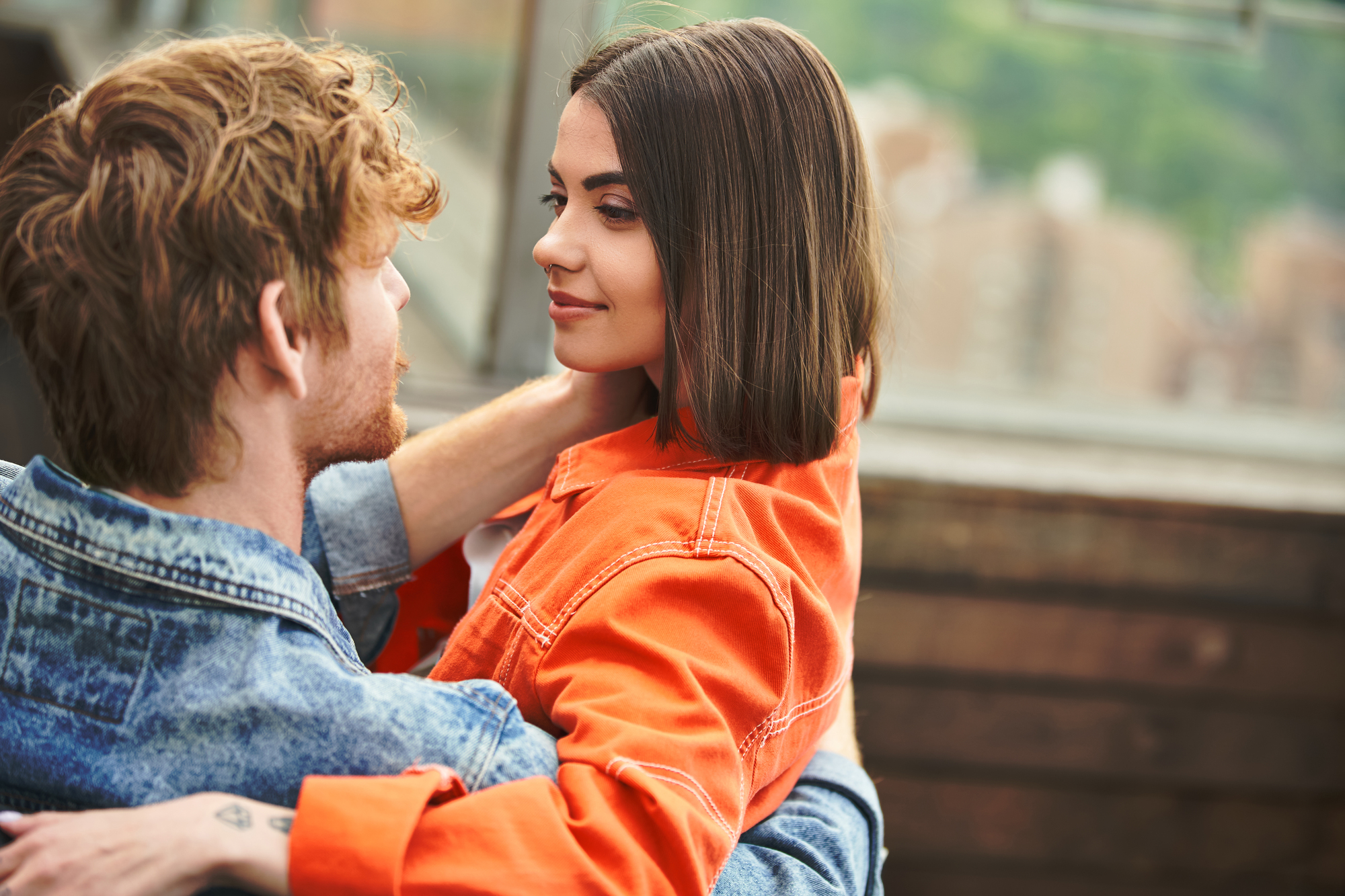 A young man and woman are sitting close together, embracing and looking into each other's eyes. The man has curly hair and a beard, wearing a denim jacket. The woman has straight shoulder-length hair and is wearing an orange jacket. A cityscape is visible in the background.